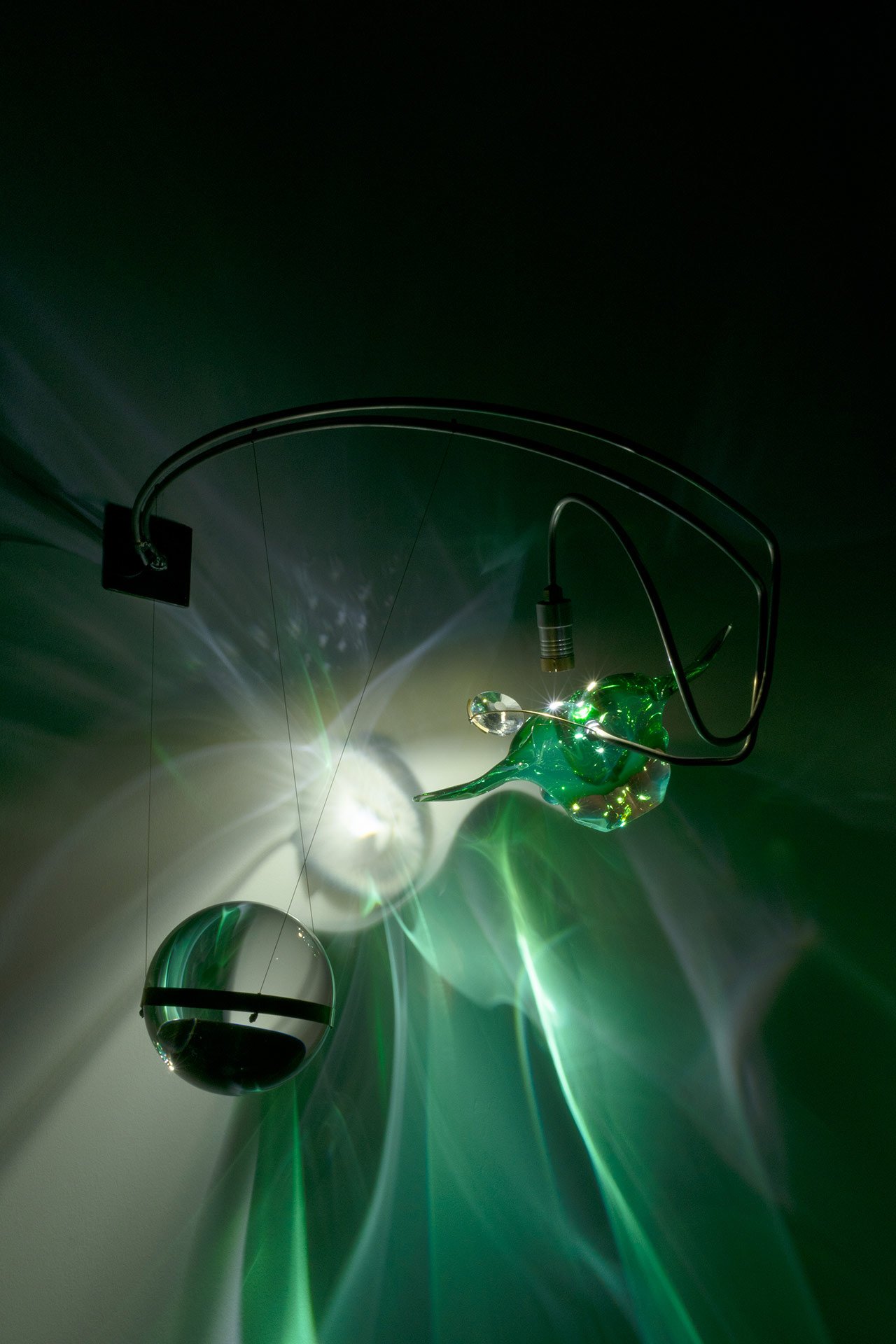 Wall-mounted fixture made of bent steel, brass accents, a green flower-shaped vintage Murano glass console bowl, and a clear glass sphere. LED-lit, it casts green and white light.