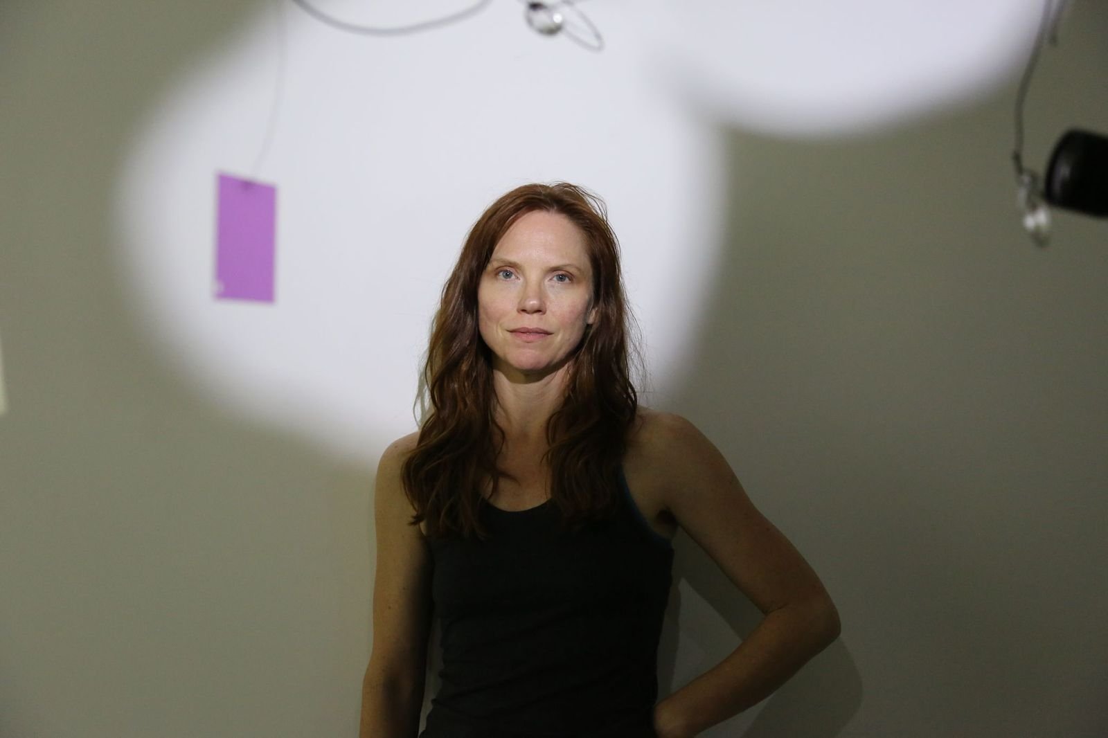 A woman with long brown hair and wearing a black tank top, standing in front of a plain wall with a lighting reflector above, during a photo shoot.