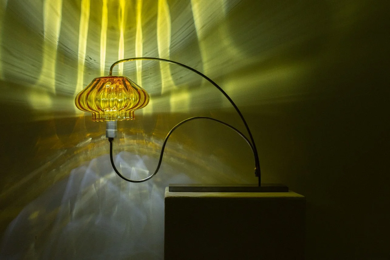 Table lamp made of bent steel piping, a steel rectangular base, brass accents, and the bottom of a yellow vintage circus tent glass jar. LED-lit, it casts yellow and white light.
