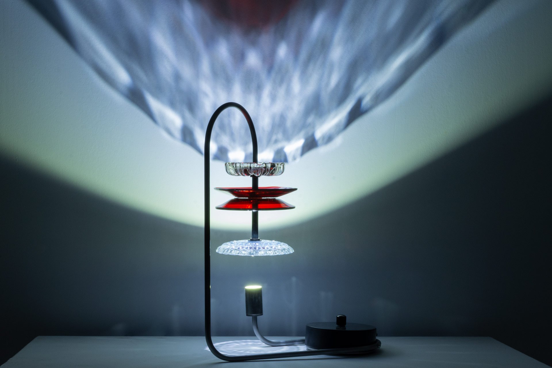Table lamp made of bent steel piping, a steel circular base, brass accents, and four vintage American glass plates (two clear, two red). LED-lit, it casts white and red light in a faceted pattern.