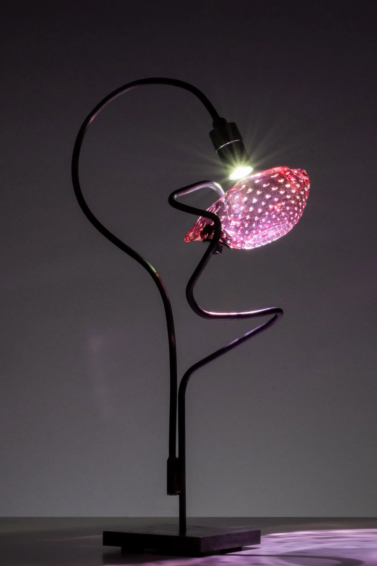 Table lamp made of bent steel piping, a steel rectangular base, brass accents, and a pink vintage bullicante glass ashtray. LED-lit, it casts pink and white light.