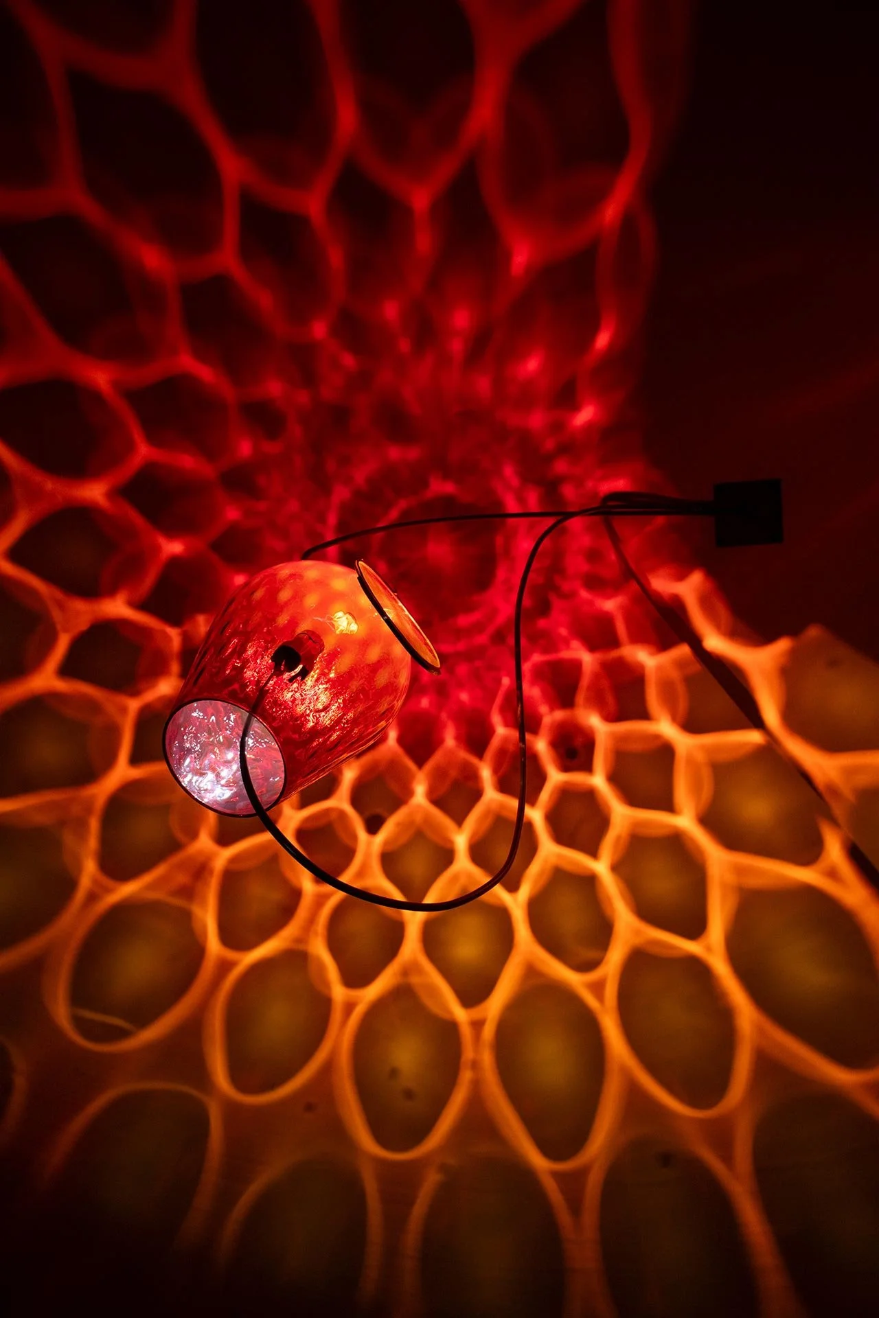 Wall-mounted fixture made of bent steel, brass accents, and a red vintage Empoli glass vase. LED-lit, it casts red and orange light in a mandala-like pattern.