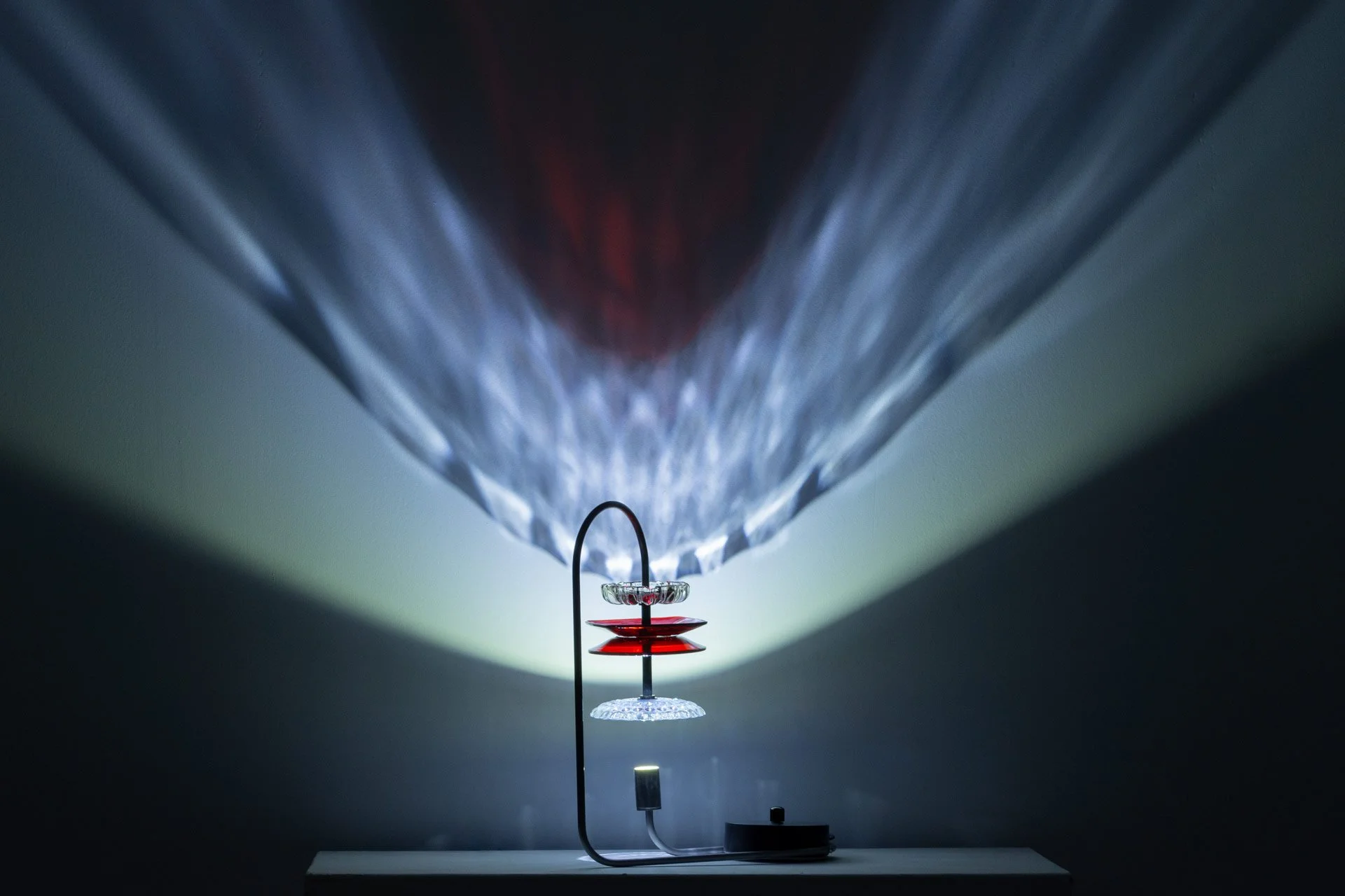 Illuminated, wider view of the lamp on a table displays white and red light in a faceted pattern.