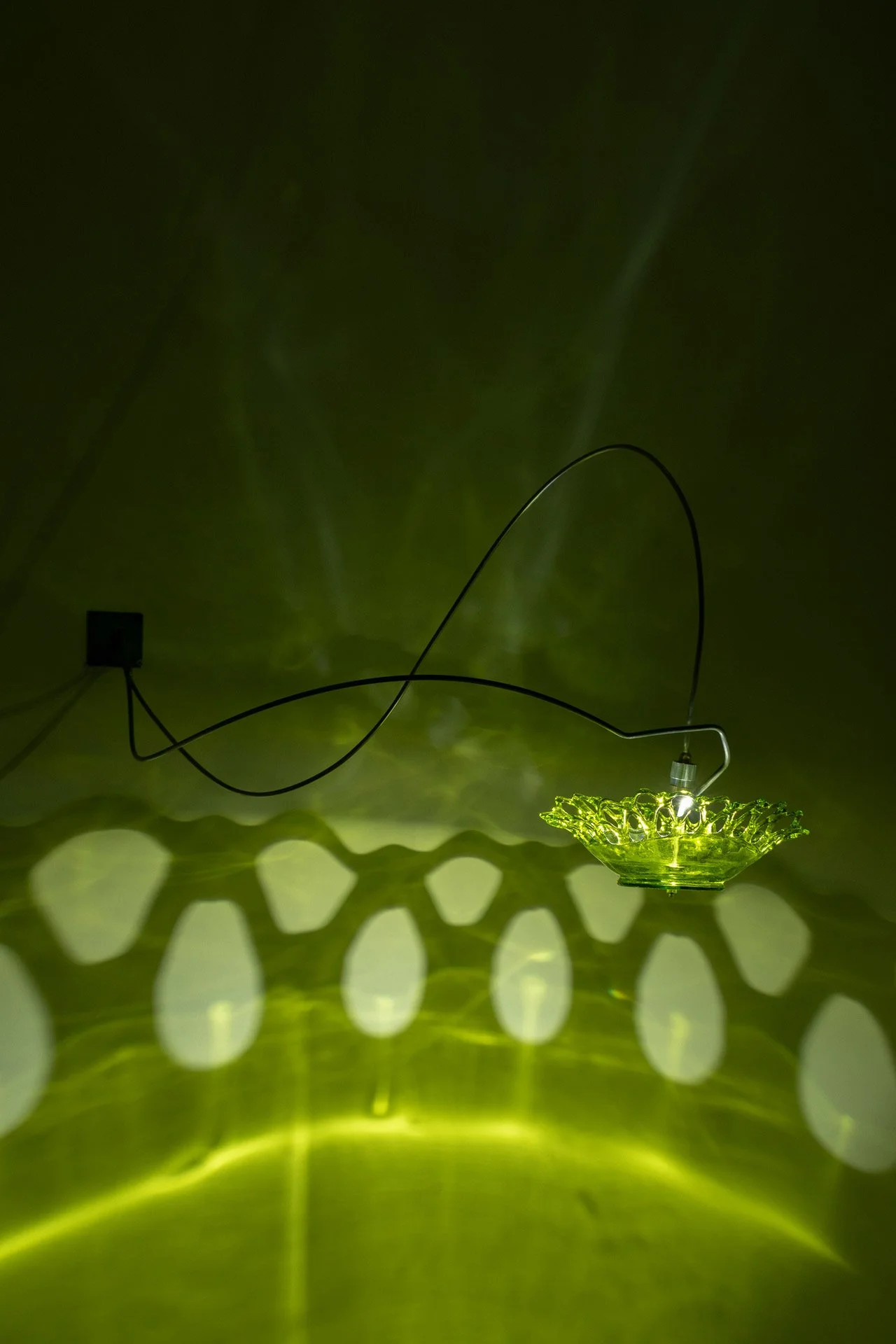 Wall-mounted fixture made of bent steel, brass accents, and a green vintage mid-century glass bowl with a lace edge. LED-lit, it casts green light with a lace pattern.