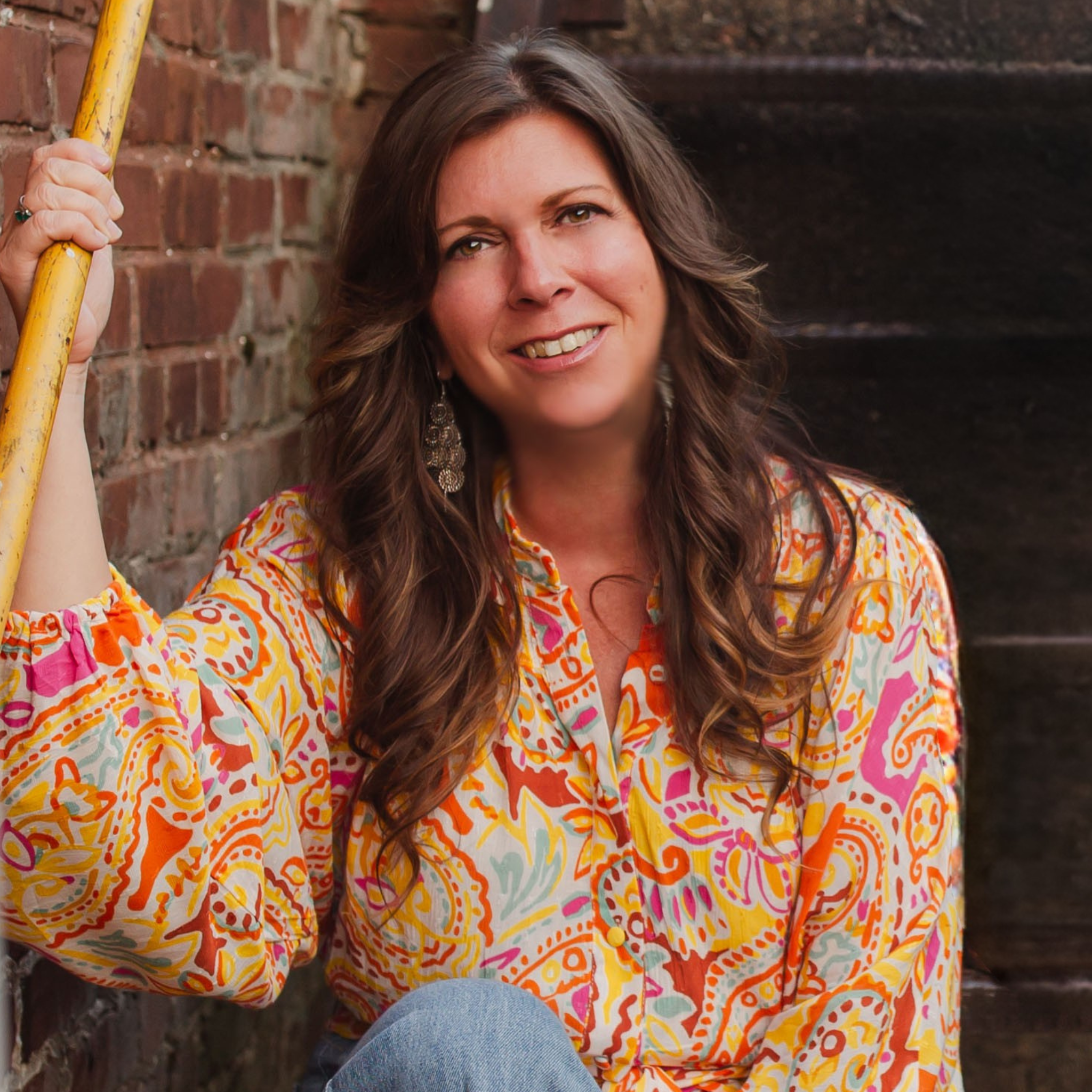 Portrait of a woman with long, wavy brown hair wearing a colorful, patterned blouse, holding a yellow handrail, sitting against a brick wall.