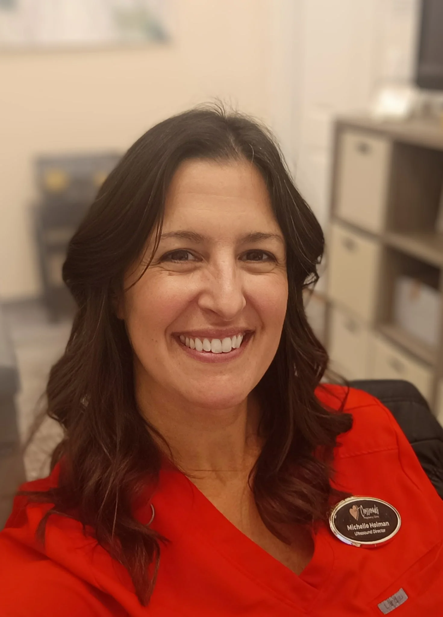 Woman smiling with brown hair, wearing a red uniform with black and white name tag, in an indoor setting with shelves and filing cabinets in the background.