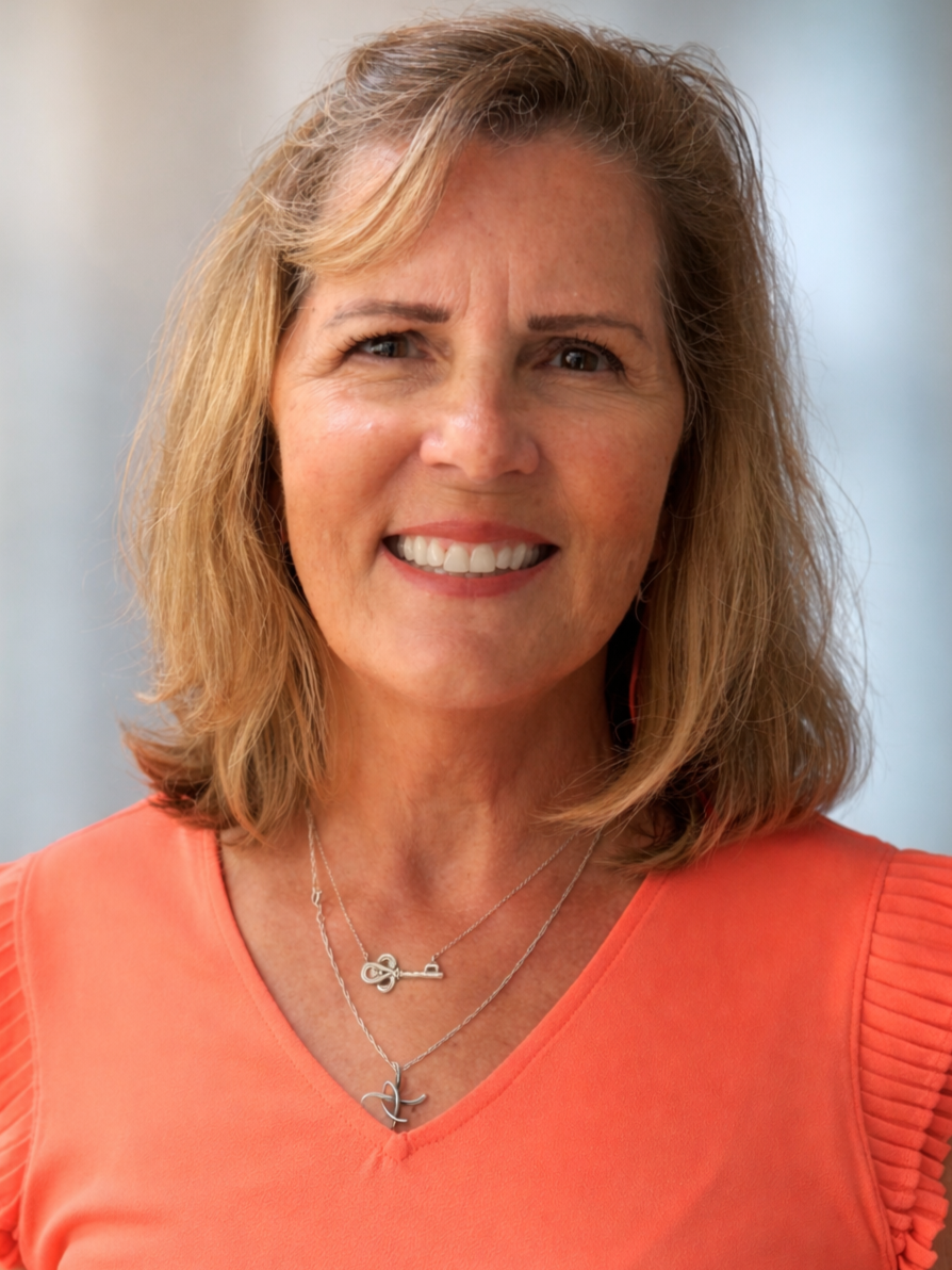 A middle-aged woman with blonde hair smiling at the camera, wearing an orange top and two silver necklaces.