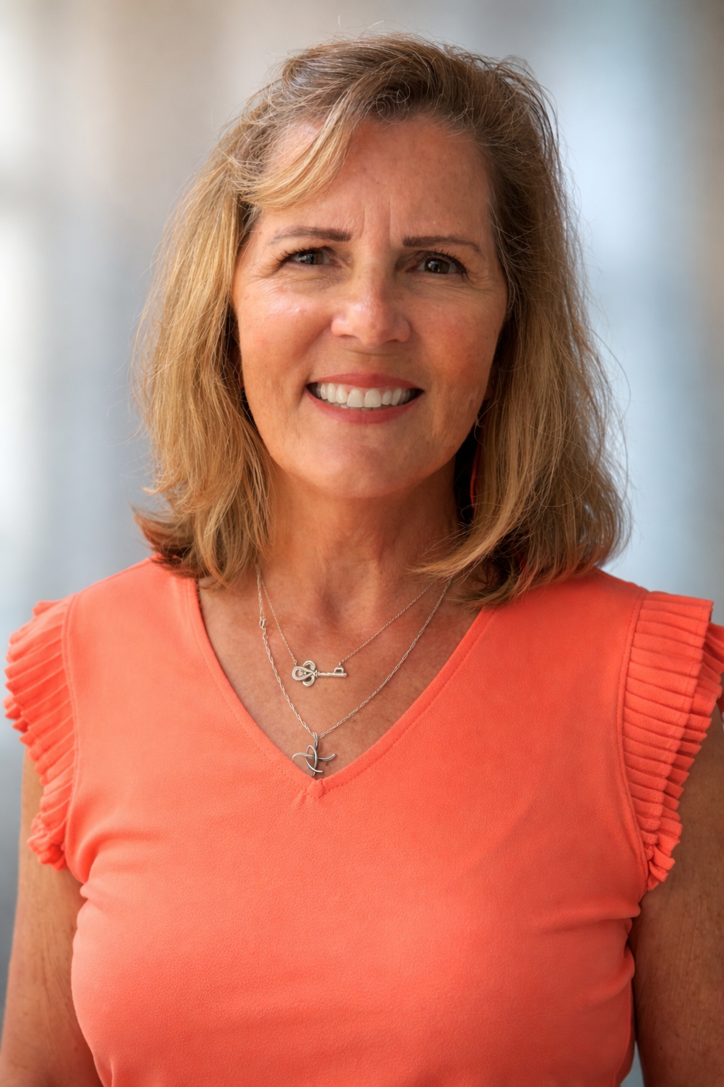 A middle-aged woman with shoulder-length blonde hair is smiling and wearing an orange sleeveless top with pleated details on the shoulders. She has two silver necklaces, one with a key pendant and the other with a bird pendant, and is standing in a softly blurred background.