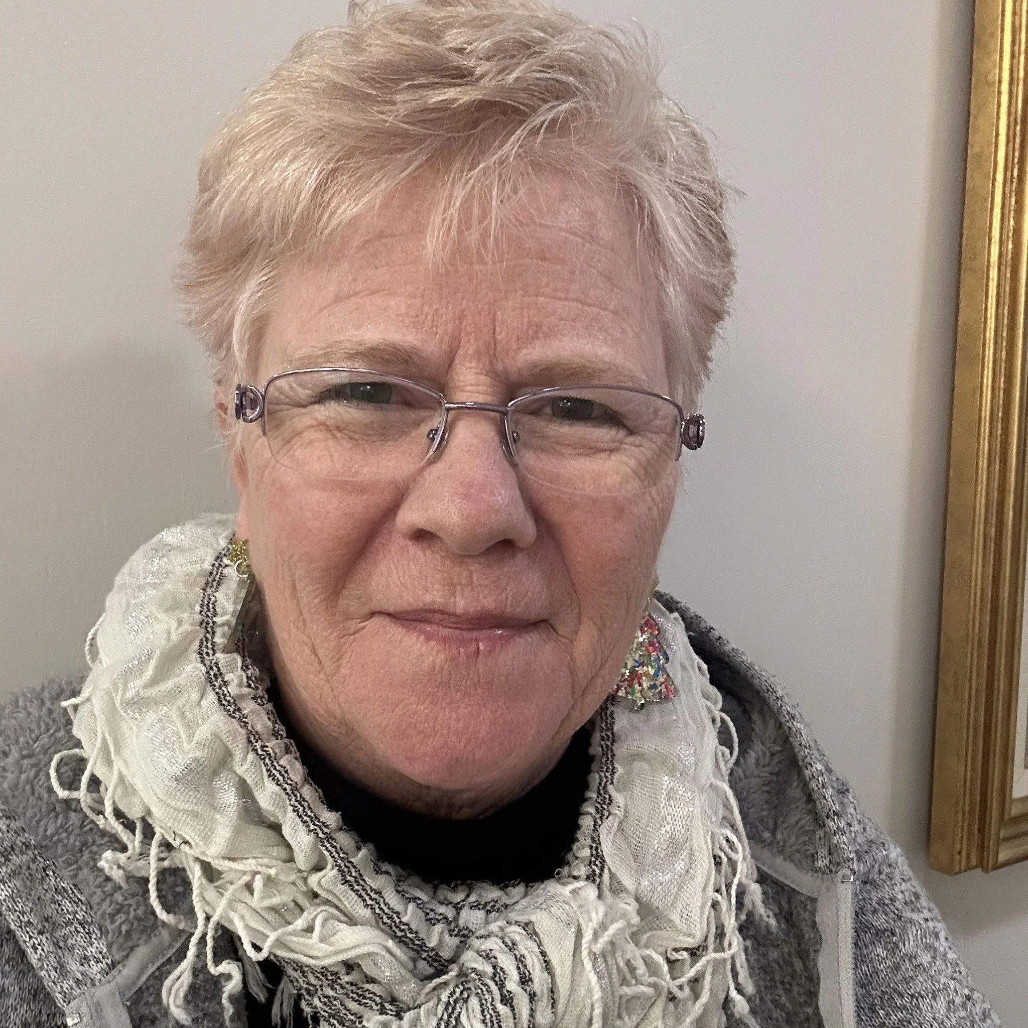A close-up of an elderly woman with short, wavy, gray hair and glasses. She is wearing a white crocheted shawl and a black top, and is smiling slightly. Part of her face and shoulders are visible, with a background that includes a gray textured surface and a gold-framed mirror or picture frame to her right.