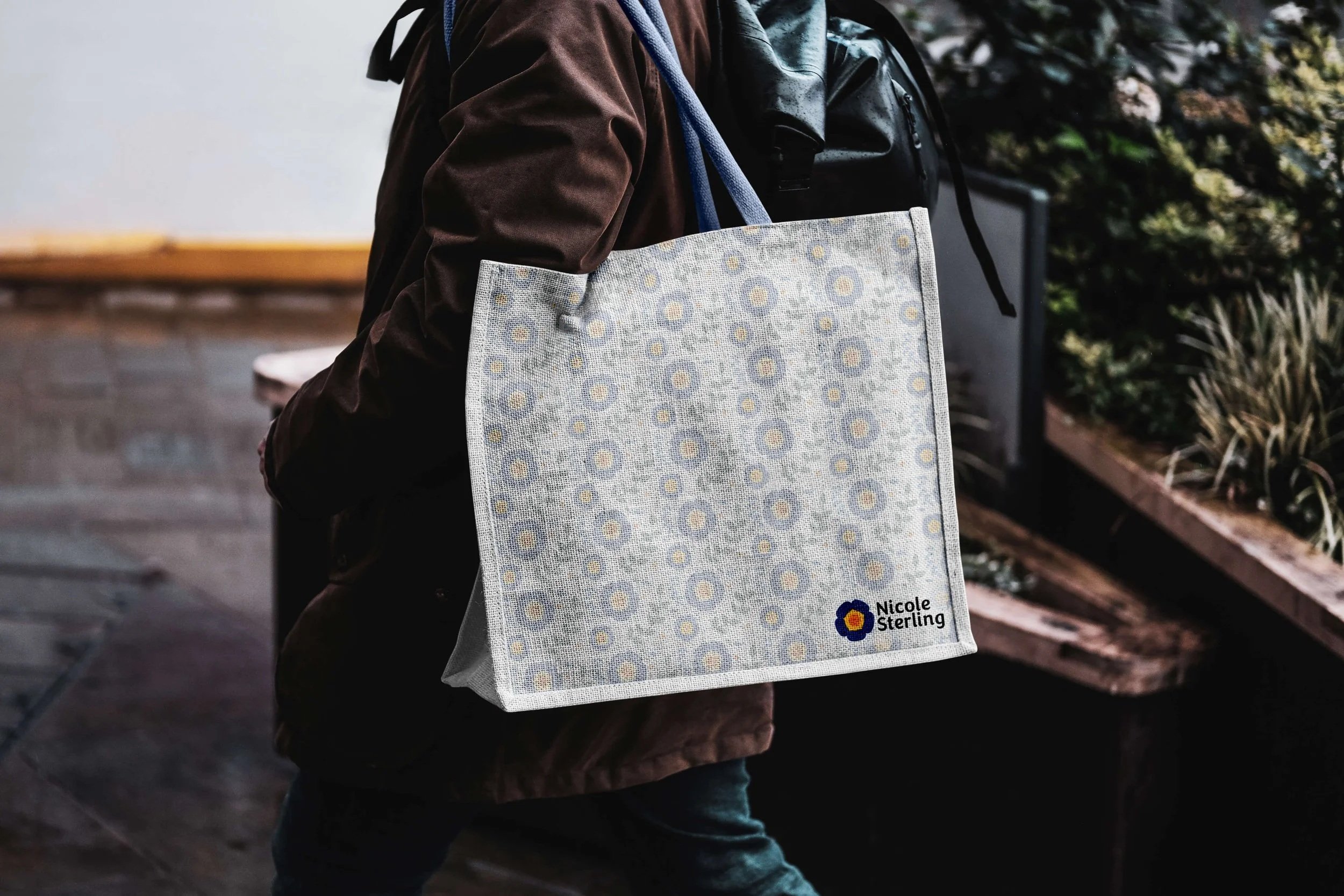 A person in a brown jacket carries a fabric tote bag with a floral pattern and the name "Nicole Sterling" embroidered on it, walking indoors near some plants.