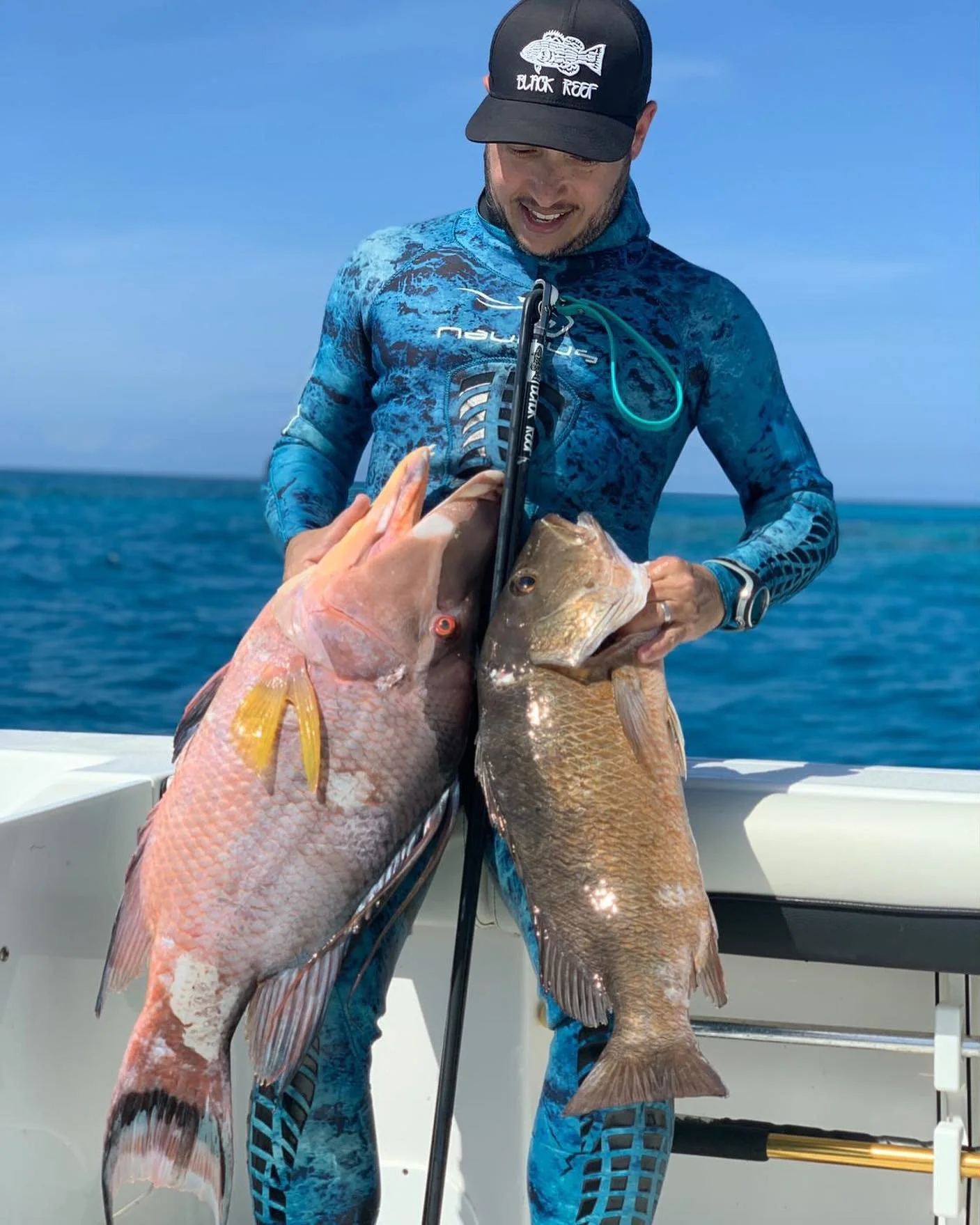 A man in blue fishing gear holding two large fish on a boat against the ocean and clear sky.