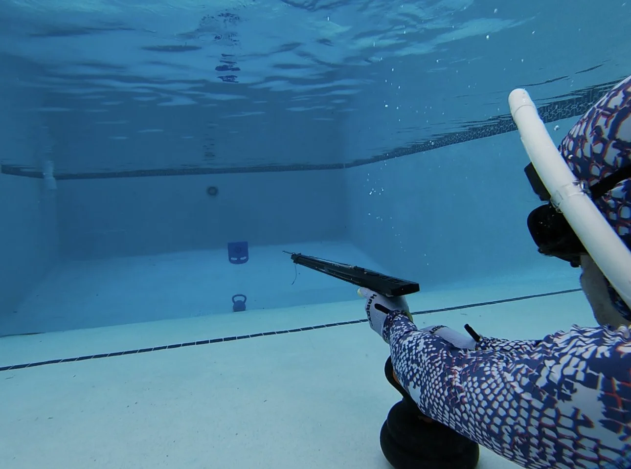 Underwater scene showing a person in diving gear aiming a spear gun at a mask and snorkel on the pool floor.
