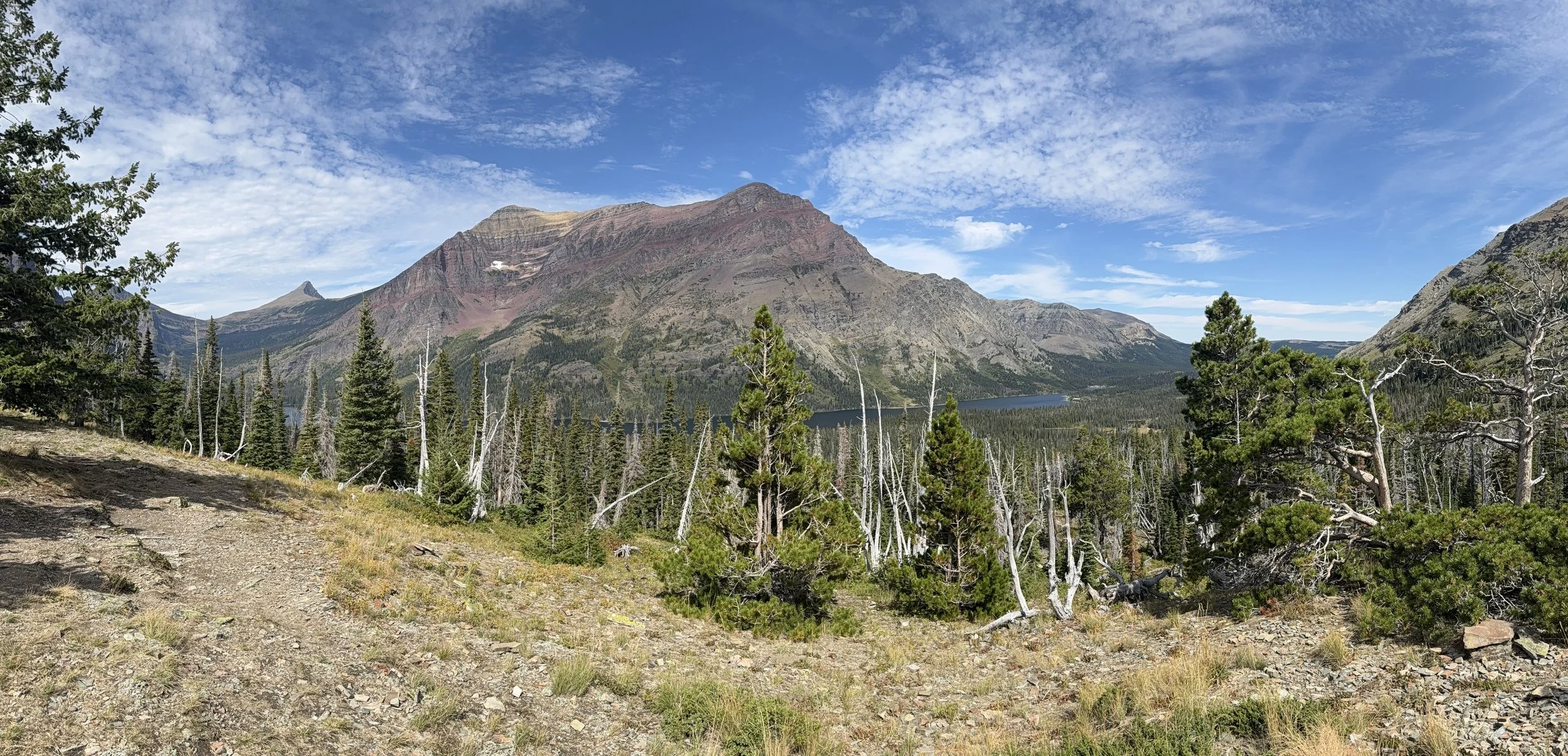 A scenic view of a mountain range with a lake at the base, surrounded by forested trees and a clear blue sky with scattered clouds.