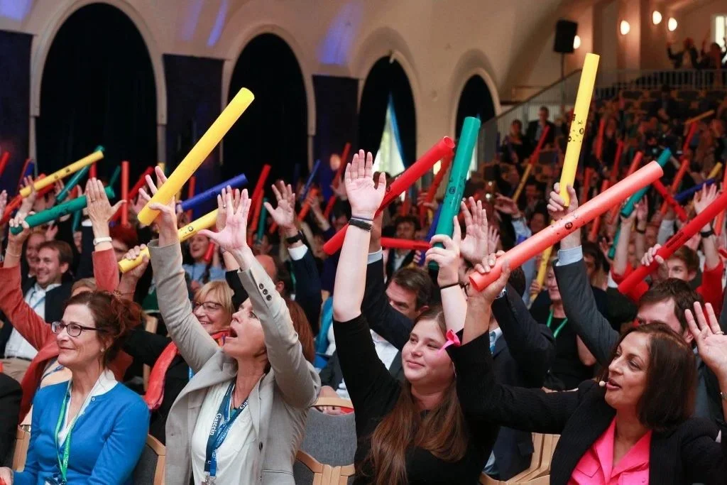 People in a conference hall raising colorful foam sticks, engaging in an energetic activity.