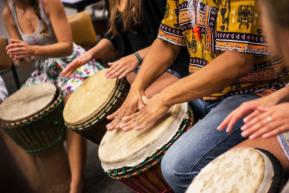 Group of people playing African drums in a workshop, with focus on their hands and colorful drum skins.