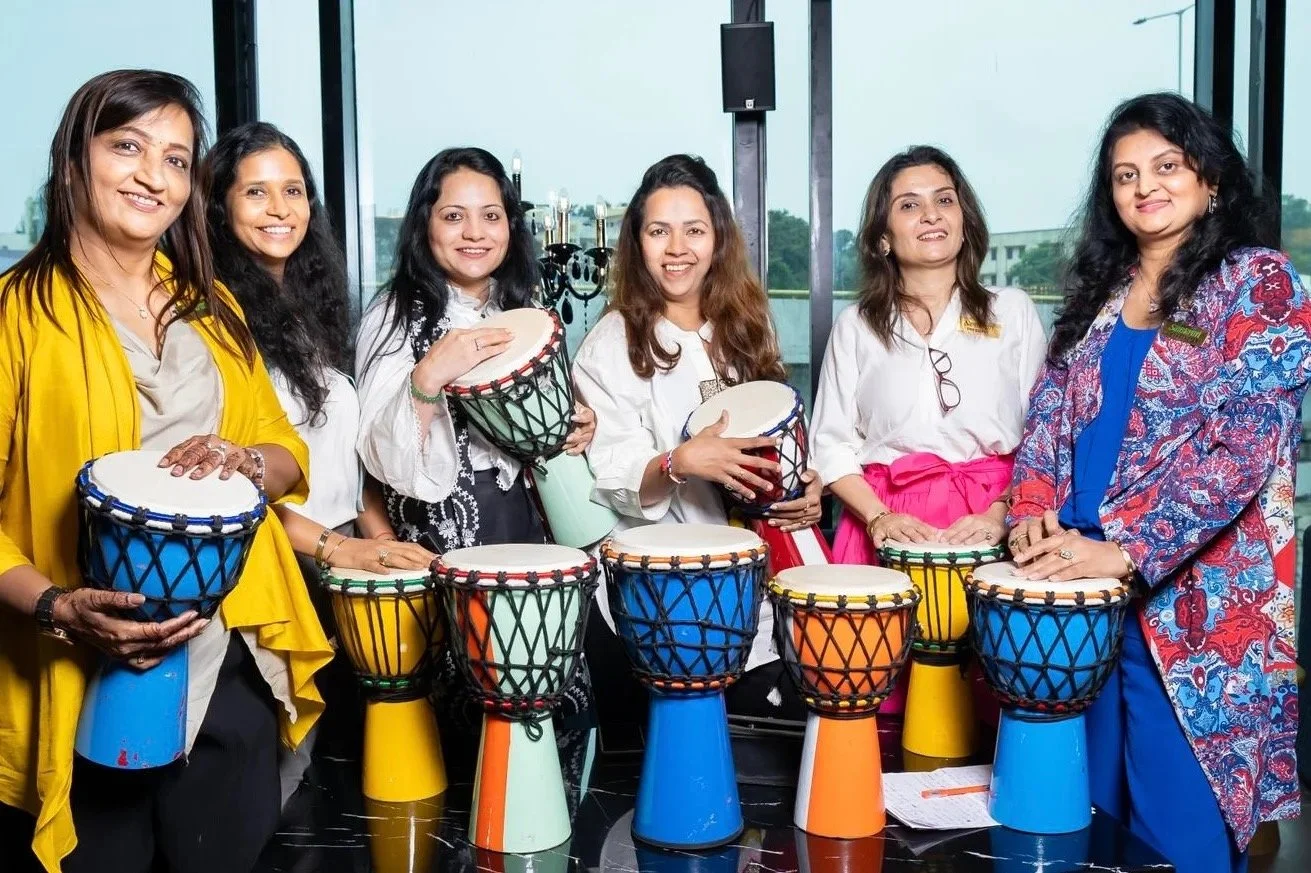 Group of women standing behind colorful drums, smiling, indoors with large windows showing outdoor scenery.
