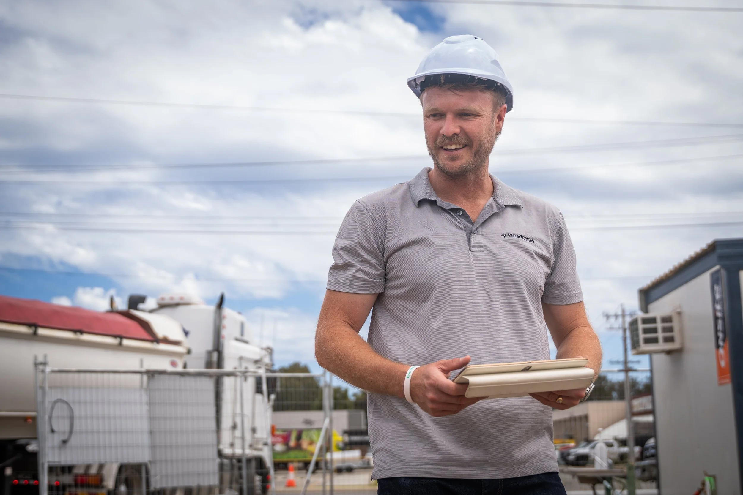 A man wearing a white safety helmet and a gray polo shirt holding a clipboard outdoors with trucks and a fence in the background.