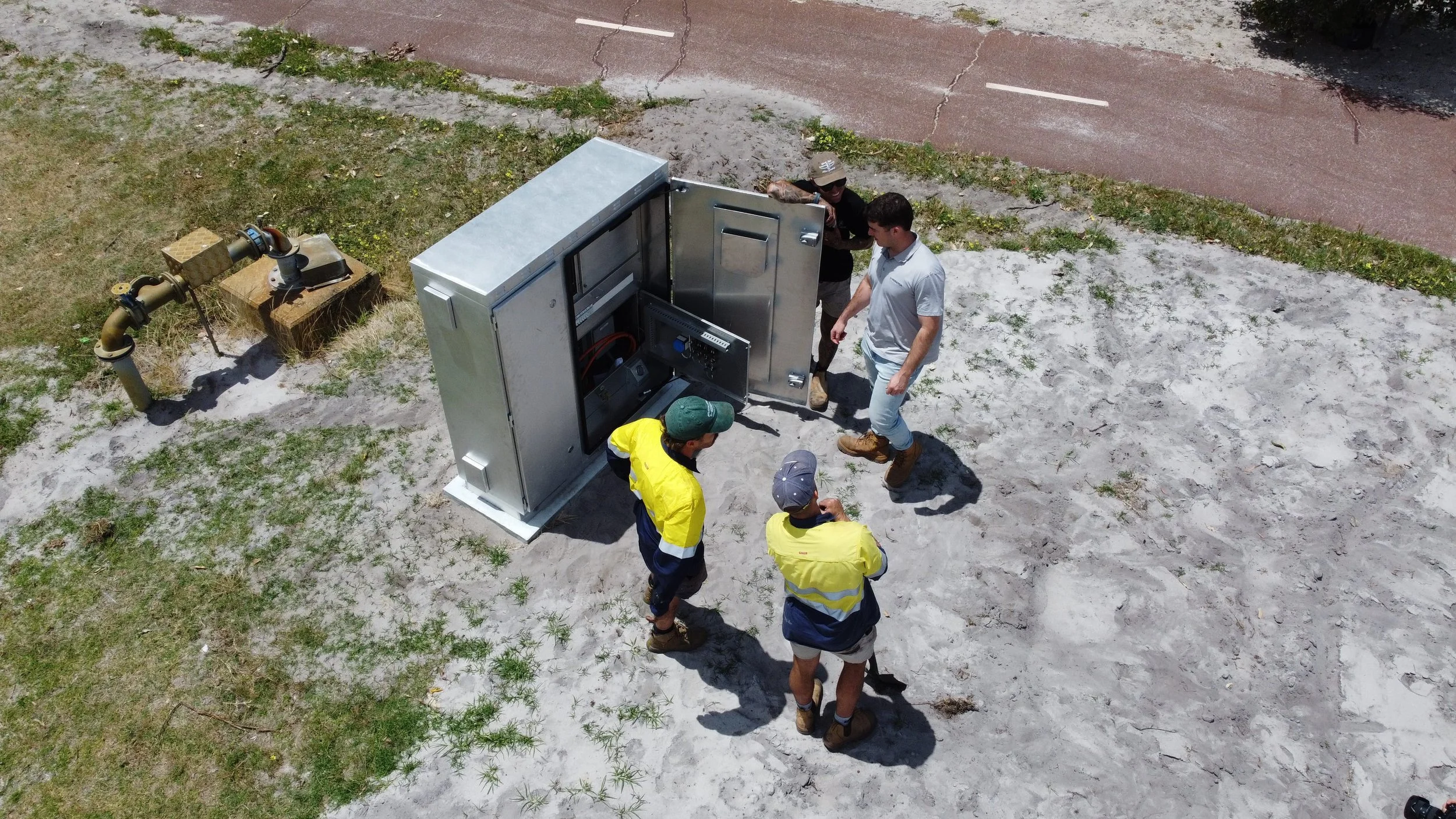 Group of four men inspecting or repairing electrical or communication equipment outdoors near a sandy area with grass patches and a street in the background.