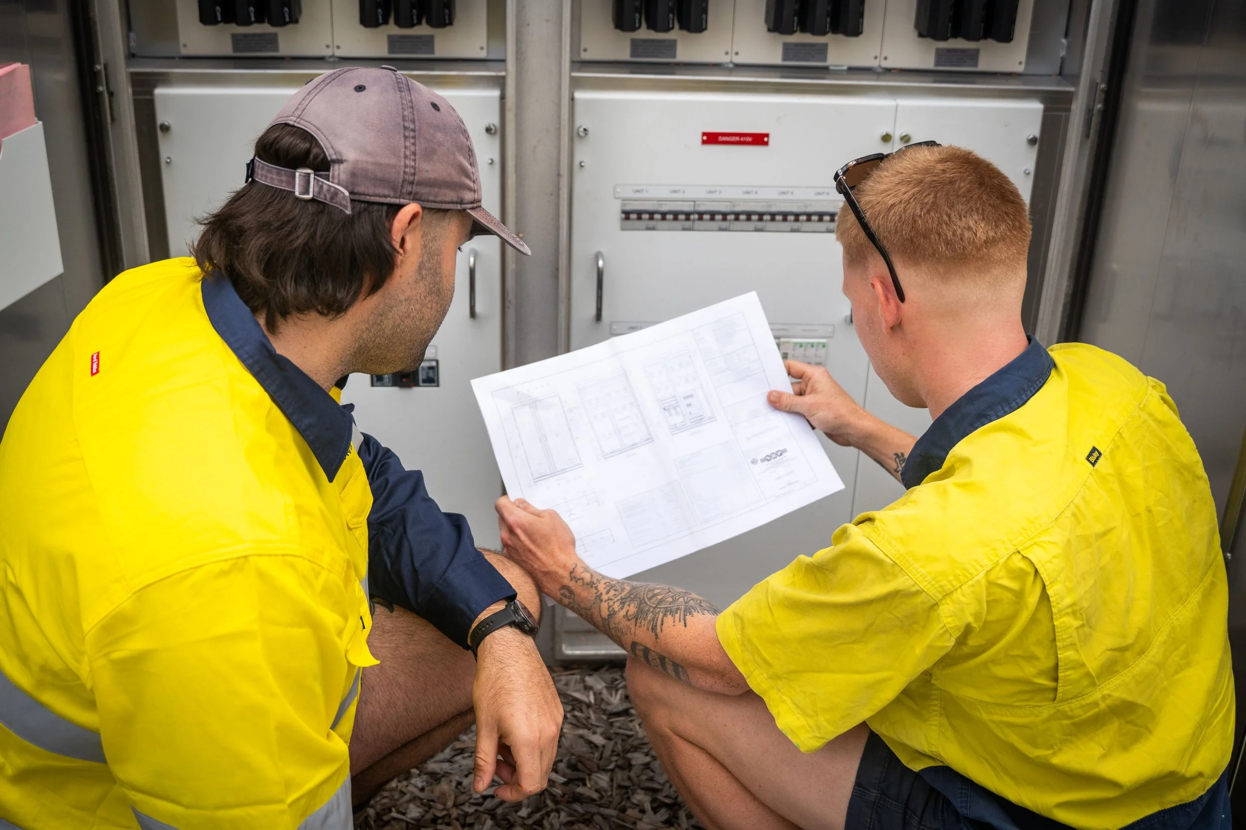 Two workers in yellow safety vests sitting on the ground reviewing electrical panels and schematics.