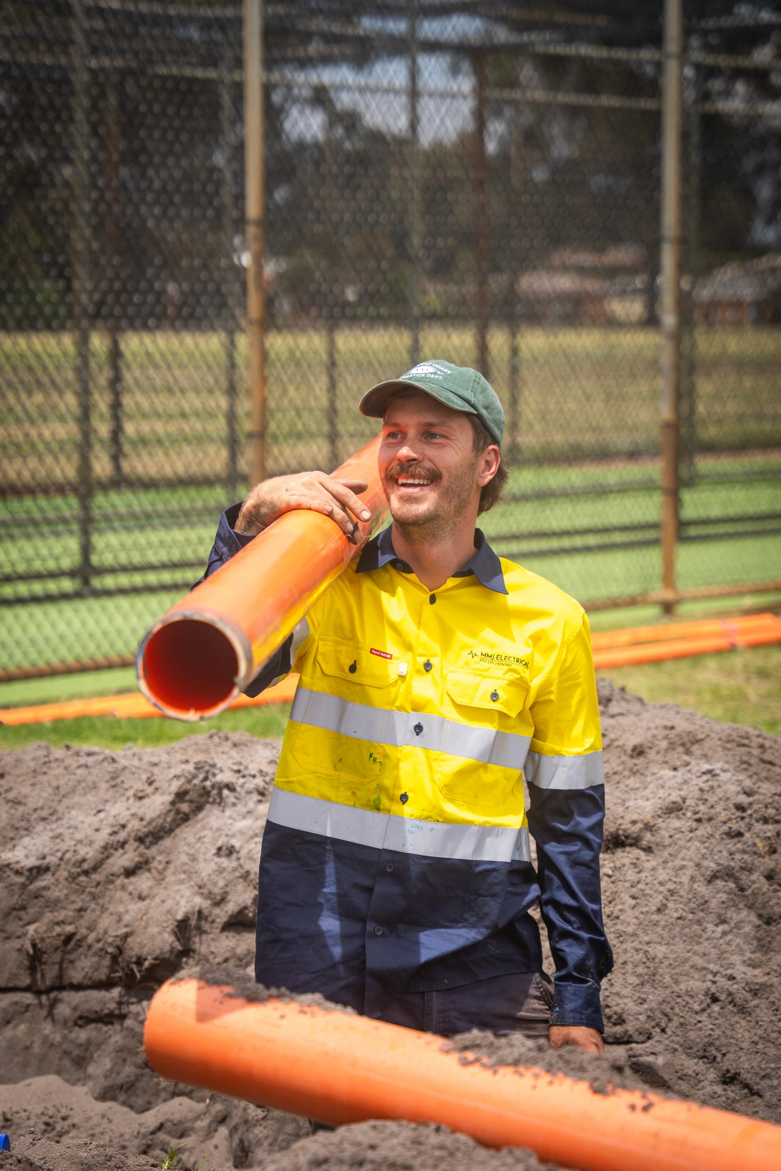 A construction worker in a yellow safety vest and green cap stands outdoors near a chain-link fence, holding an orange PVC pipe on his shoulder and smiling.