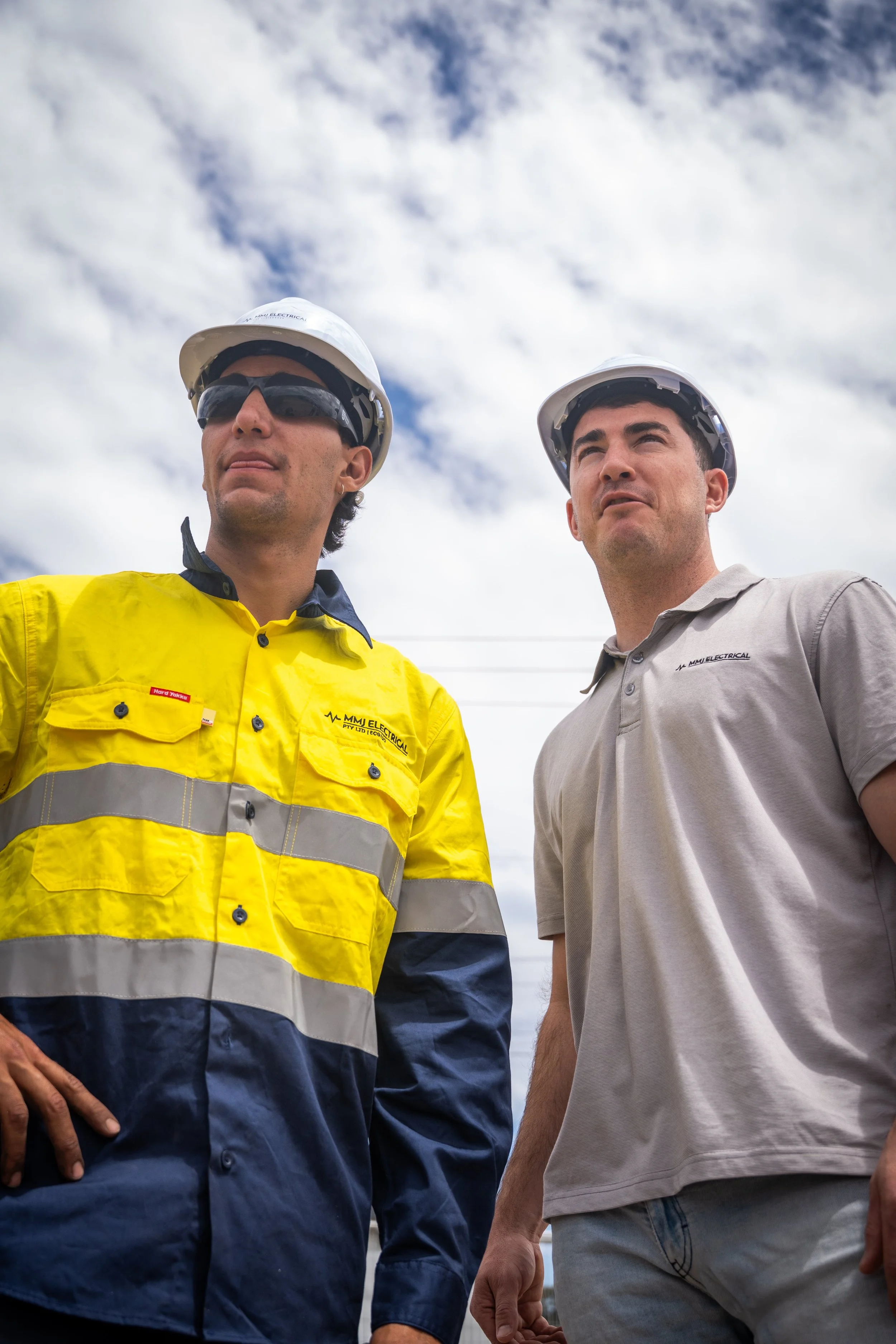 Two male utility workers wearing hard hats and safety clothes standing outdoors under a partly cloudy sky.