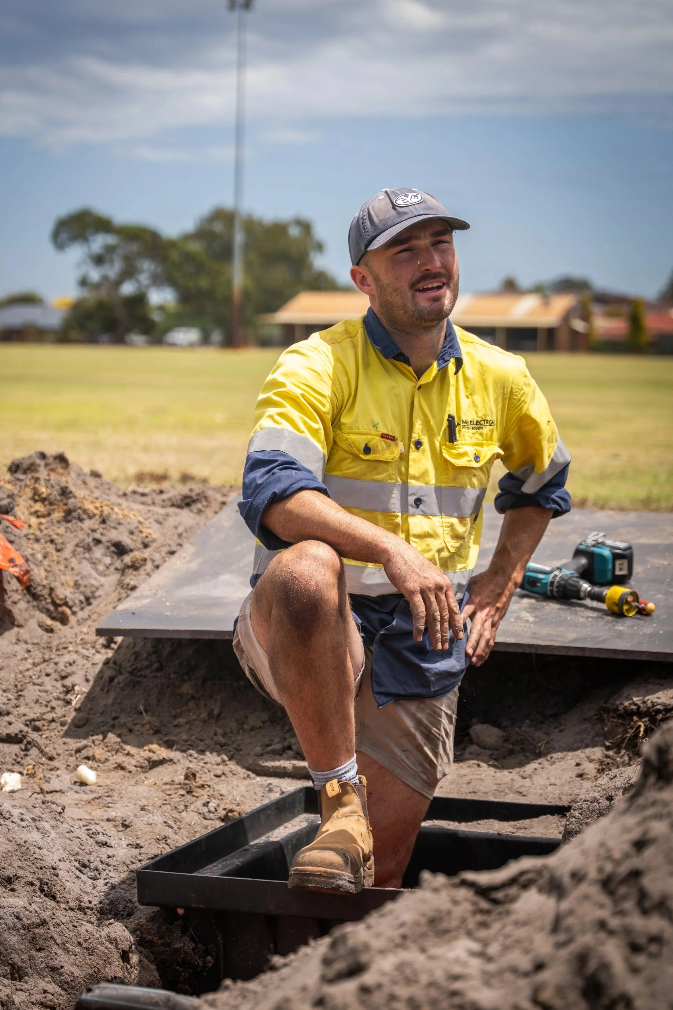 A man kneeling in a trench at an outdoor construction site, wearing a yellow safety shirt, tan shorts, and work boots, with tools nearby, under an overcast sky.