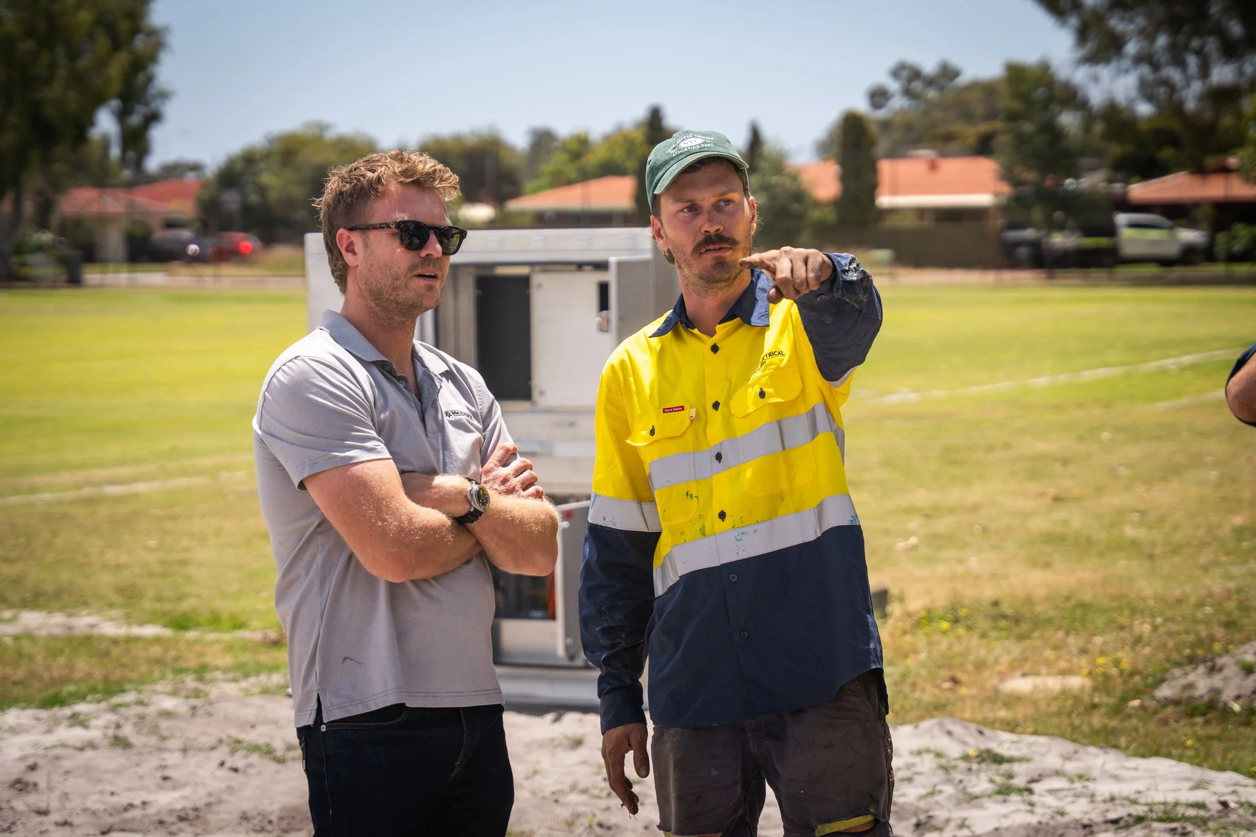 Two men standing outdoors, one in a yellow high-visibility shirt and the other in a gray polo shirt, engaged in conversation. The man in high visibility is pointing while the other listens. The background shows a grassy field and houses with trees.