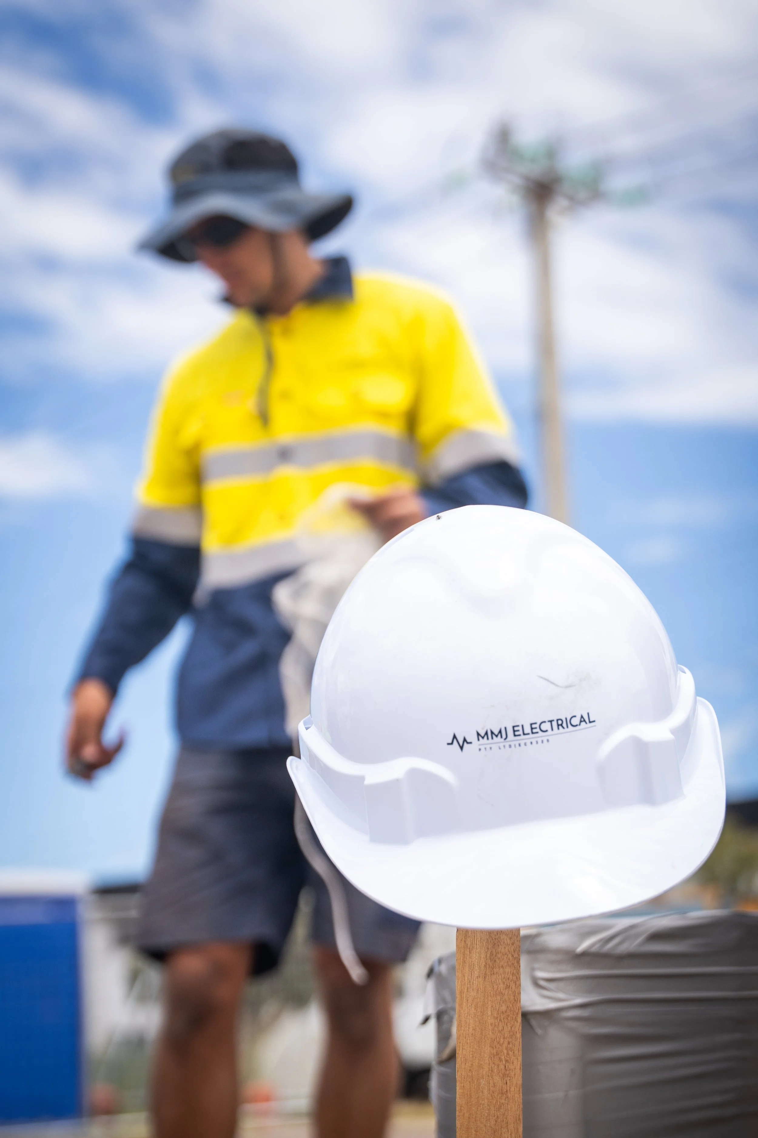 A safety helmet on a wooden post with the text 'MMJ Electrical' along with a logo, with a blurred construction worker in a yellow safety vest in the background outdoors under a partly cloudy sky.