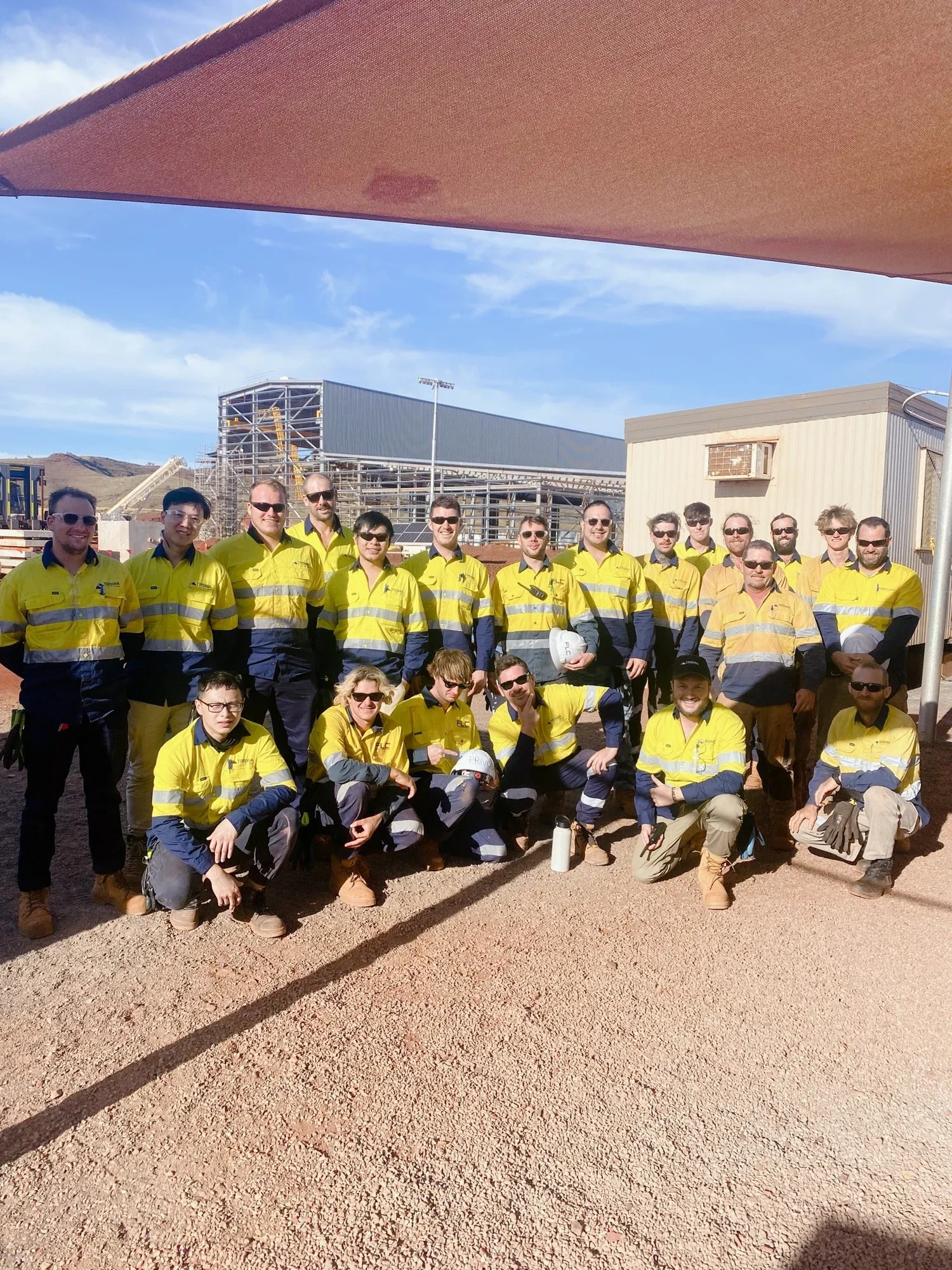 Group of workers wearing yellow and navy safety uniforms, posing outdoors at a construction site with industrial buildings in the background.