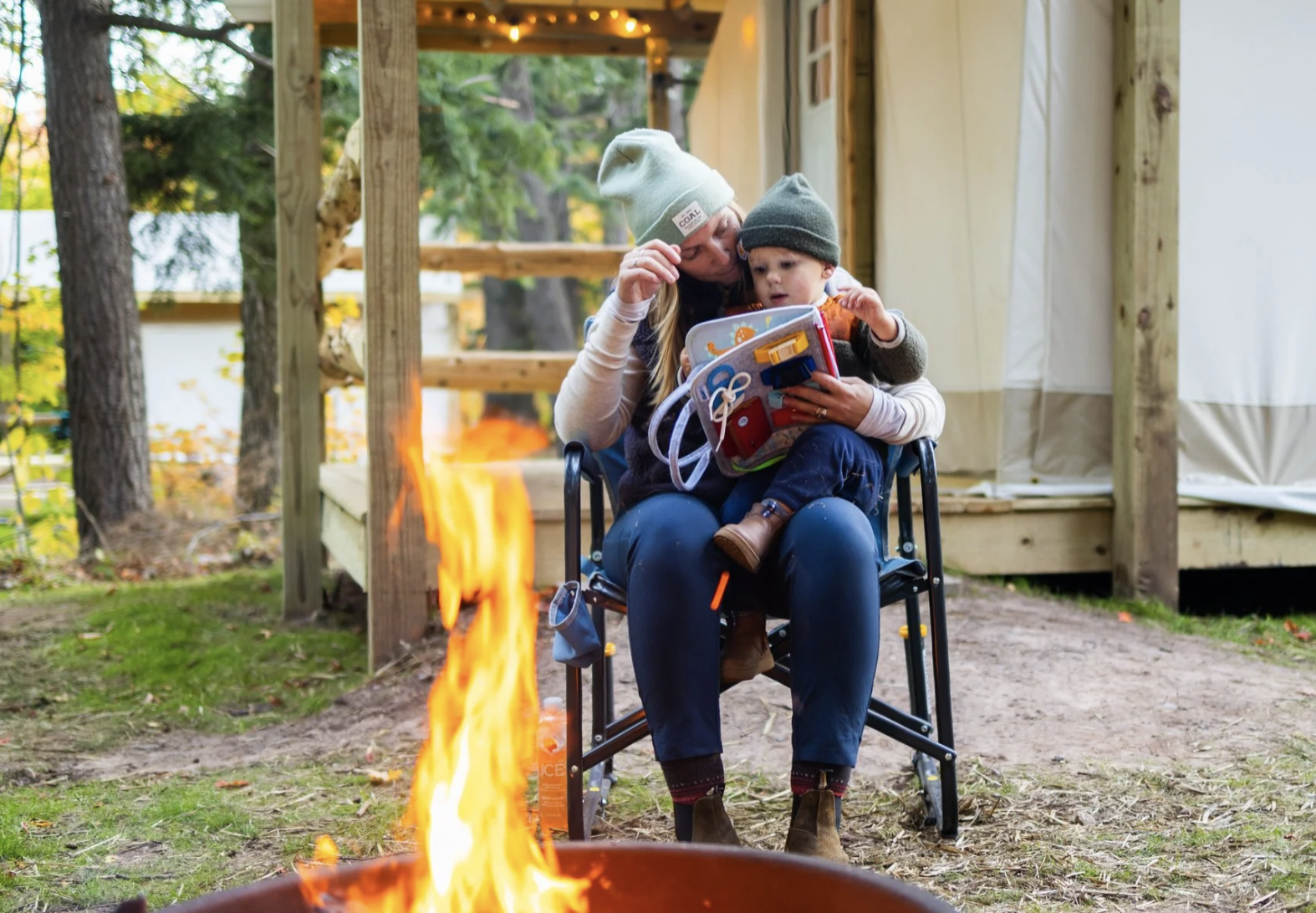 A woman and young boy sitting outside on a chair near a campfire. The woman is wearing a beige beanie and is showing the boy a small red and white toolbox. The boy is wearing a dark beanie and is looking at the toolbox. There are trees and a wooden structure in the background.