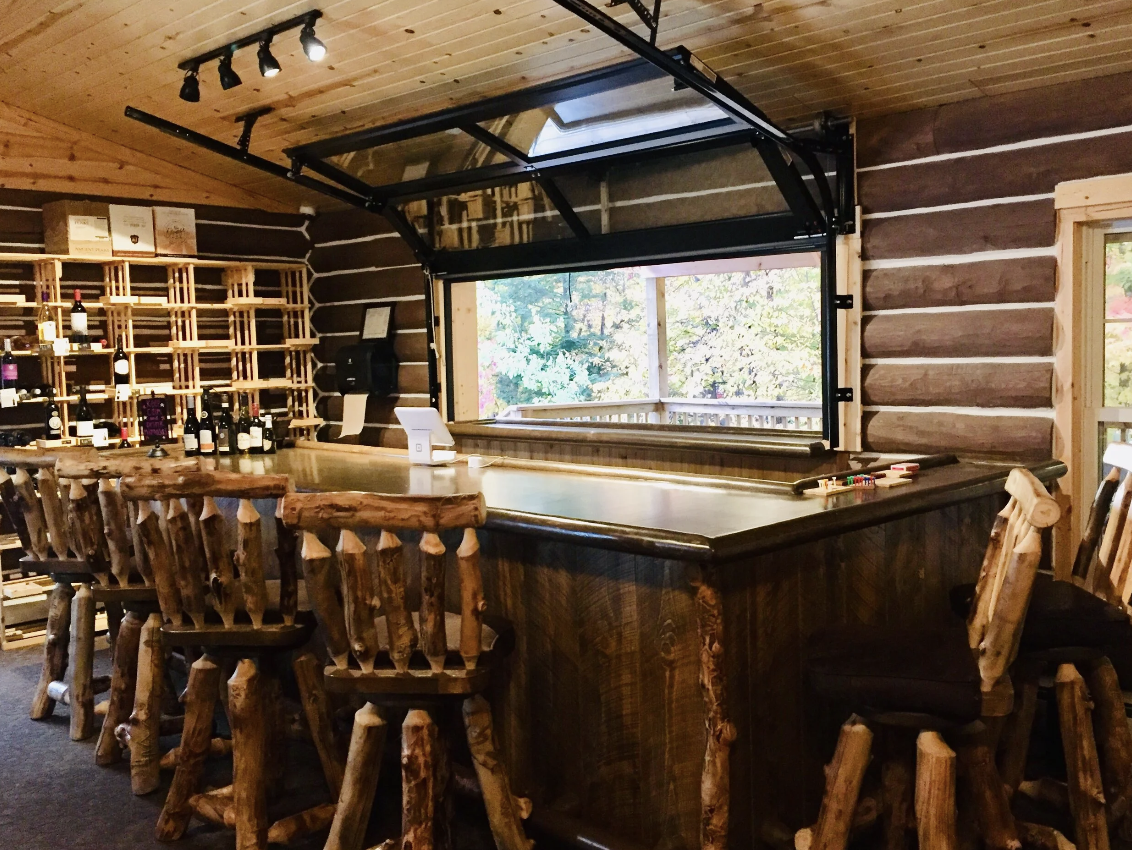 Rustic log cabin-style bar with wooden chairs, a wine display, and an open garage-style window showing trees outside.