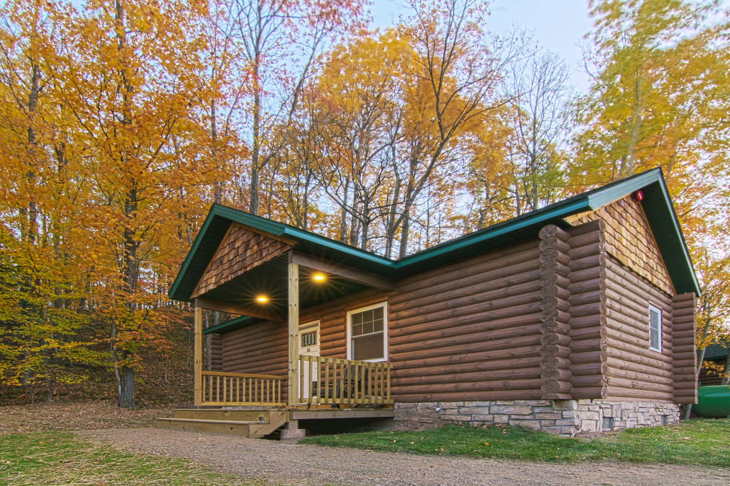 A rustic log cabin with a green metal roof and a small front porch with wooden railing, set against a backdrop of autumn trees with colorful fall foliage.