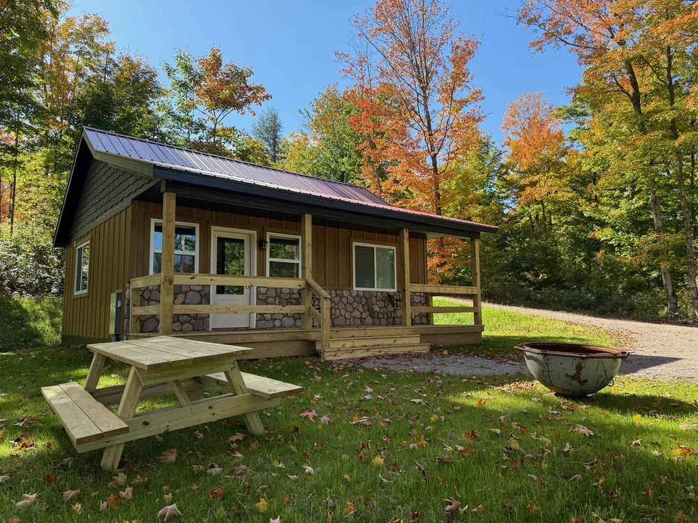 A small house with a metal roof and wooden siding, surrounded by trees with autumn foliage, has a front porch with a railing. A wooden picnic table is in the grass nearby. There is a gravel driveway on the right and a large metal fire pit or planter 