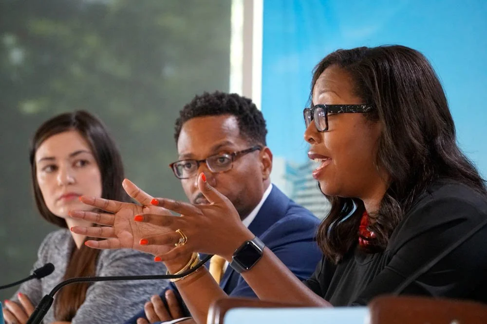 Three diverse women speaking and listening at a panel discussion, with microphones in front of them.