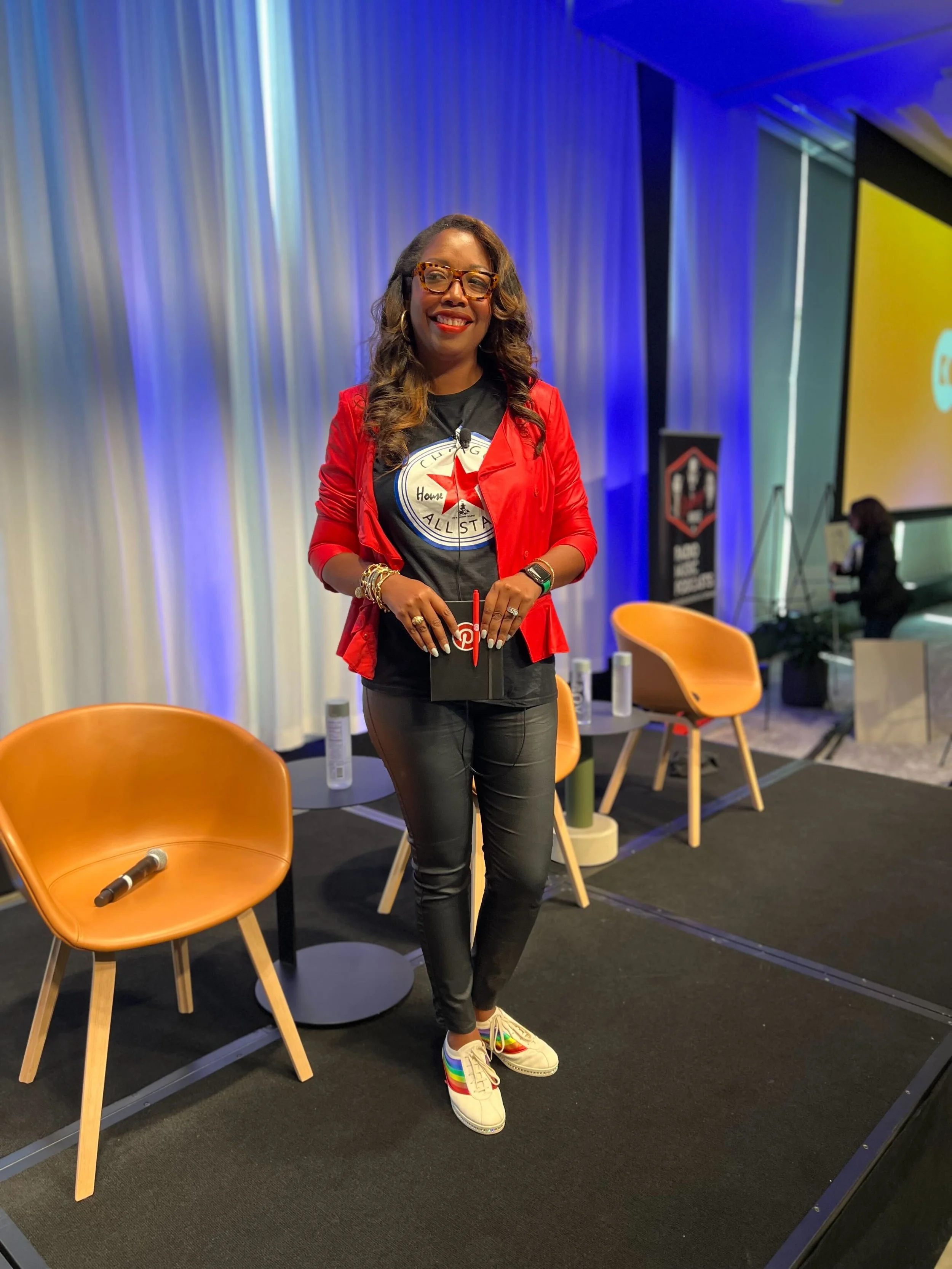 A woman wearing glasses, a red jacket, and rainbow shoelaces, standing on a stage with chairs and a large screen in the background.