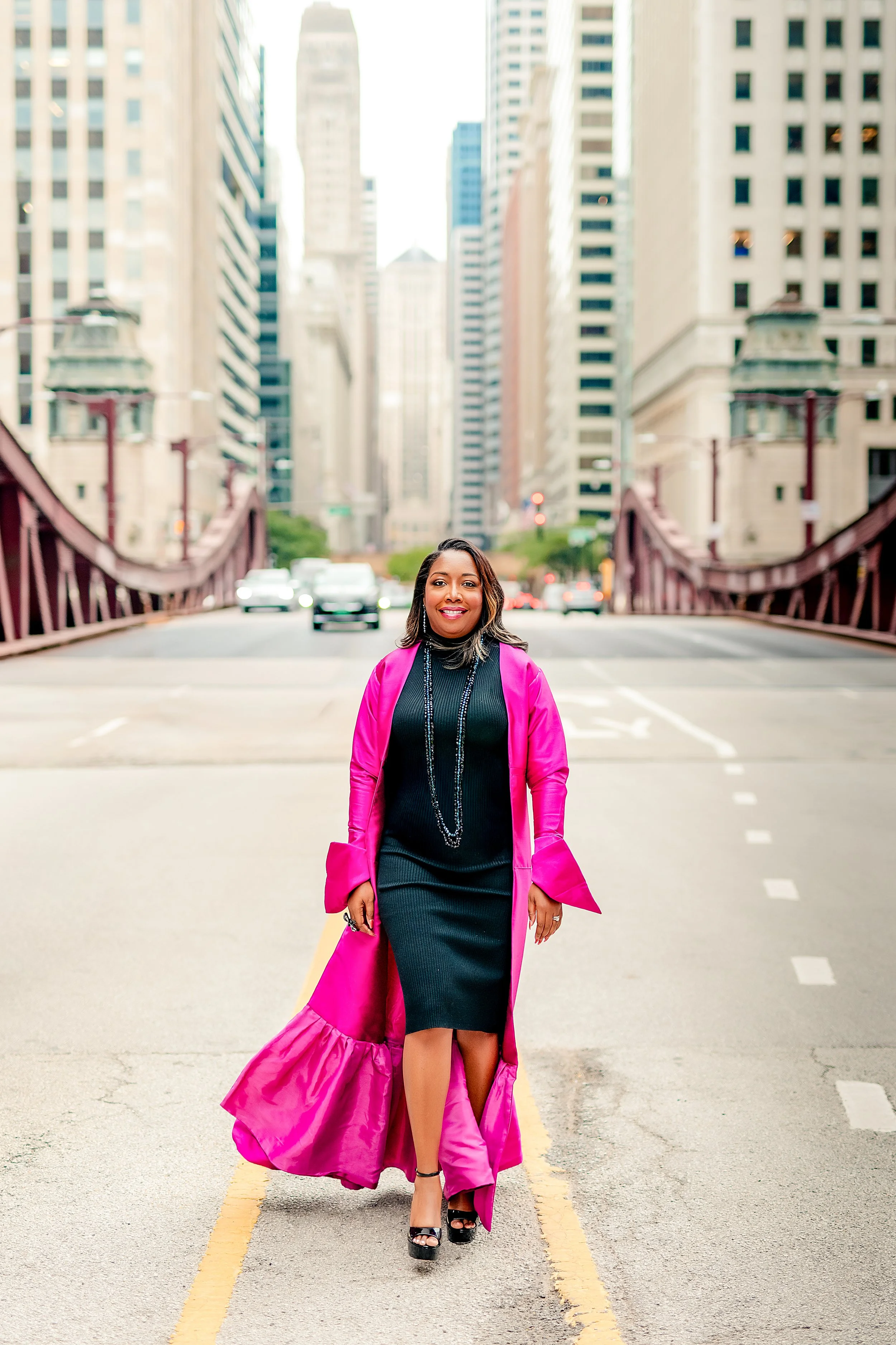 A woman standing in the middle of a city street with tall buildings in the background. She is wearing a black dress, black high heels, and a bright pink coat with ruffles at the bottom. She is smiling and looking at the camera.