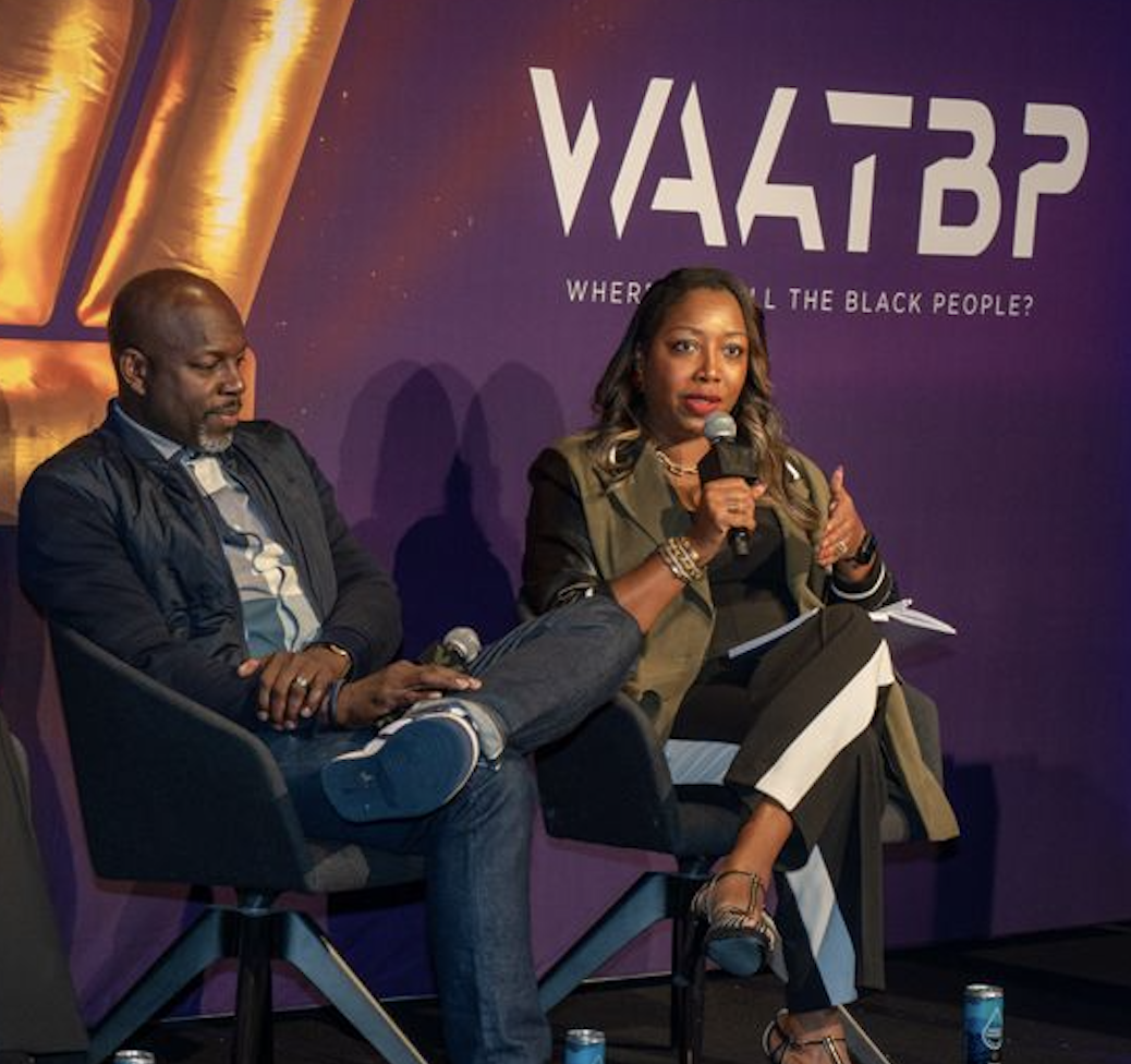 Two individuals seated on a stage, a woman speaking into a microphone and a man listening. Behind them is a purple backdrop with the text 'VBTBR' and a question mark.