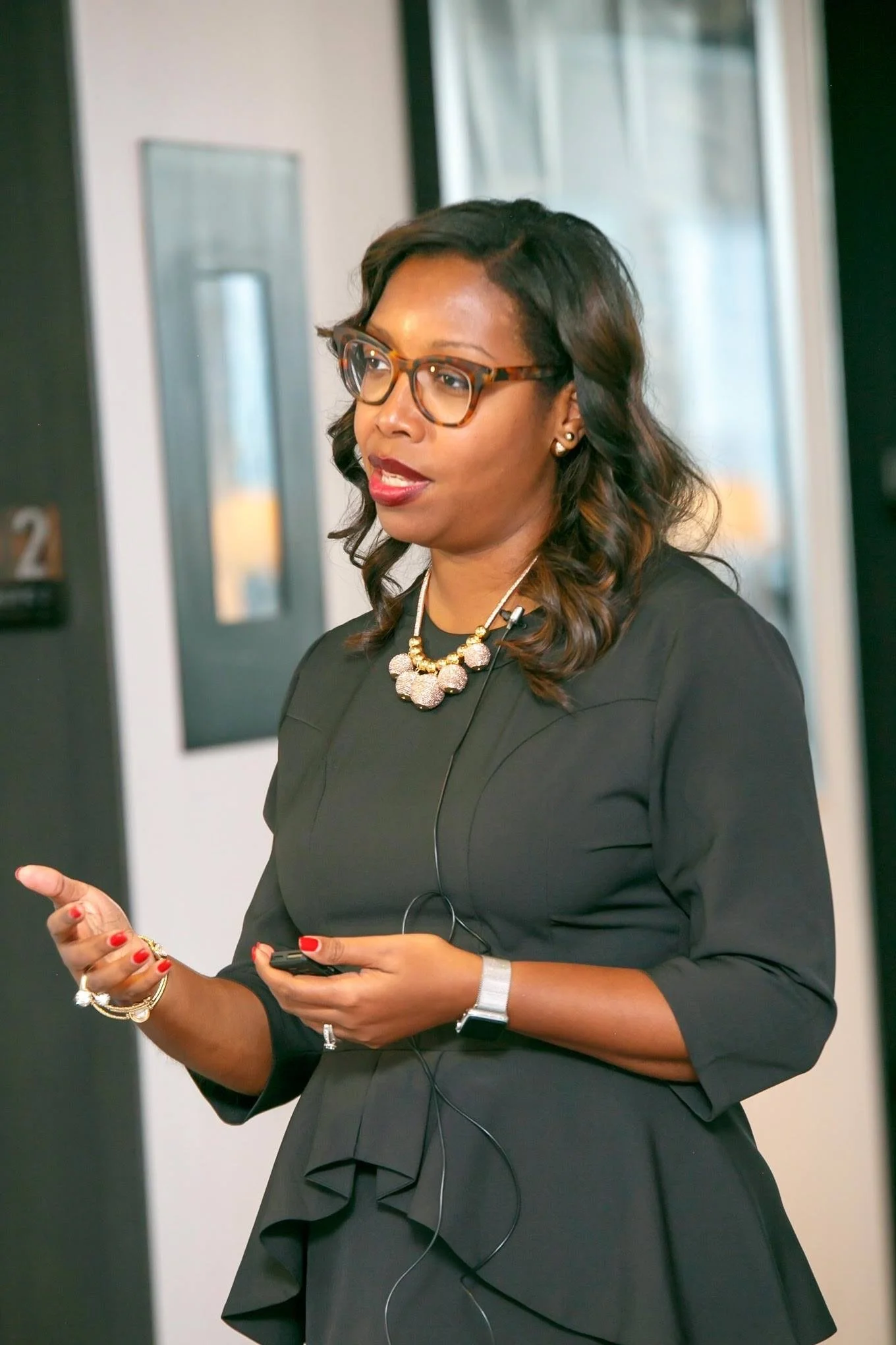 A woman with glasses and dark wavy hair, dressed in black, wearing a statement necklace and jewelry, holding a remote or pointer, speaking or presenting in an indoor setting.