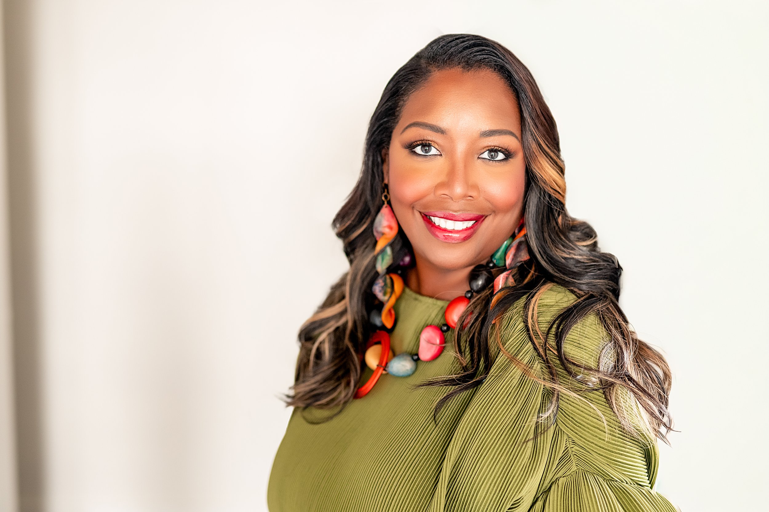 A smiling woman with dark, wavy hair wearing a green top and colorful statement necklace and earrings.