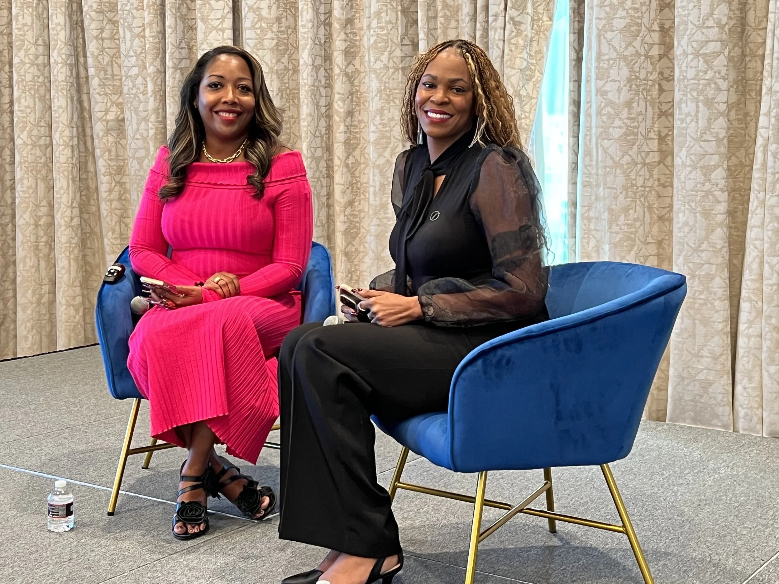 Two women sitting on blue chairs during an indoor event, smiling, with beige curtains and a window with curtains behind them.