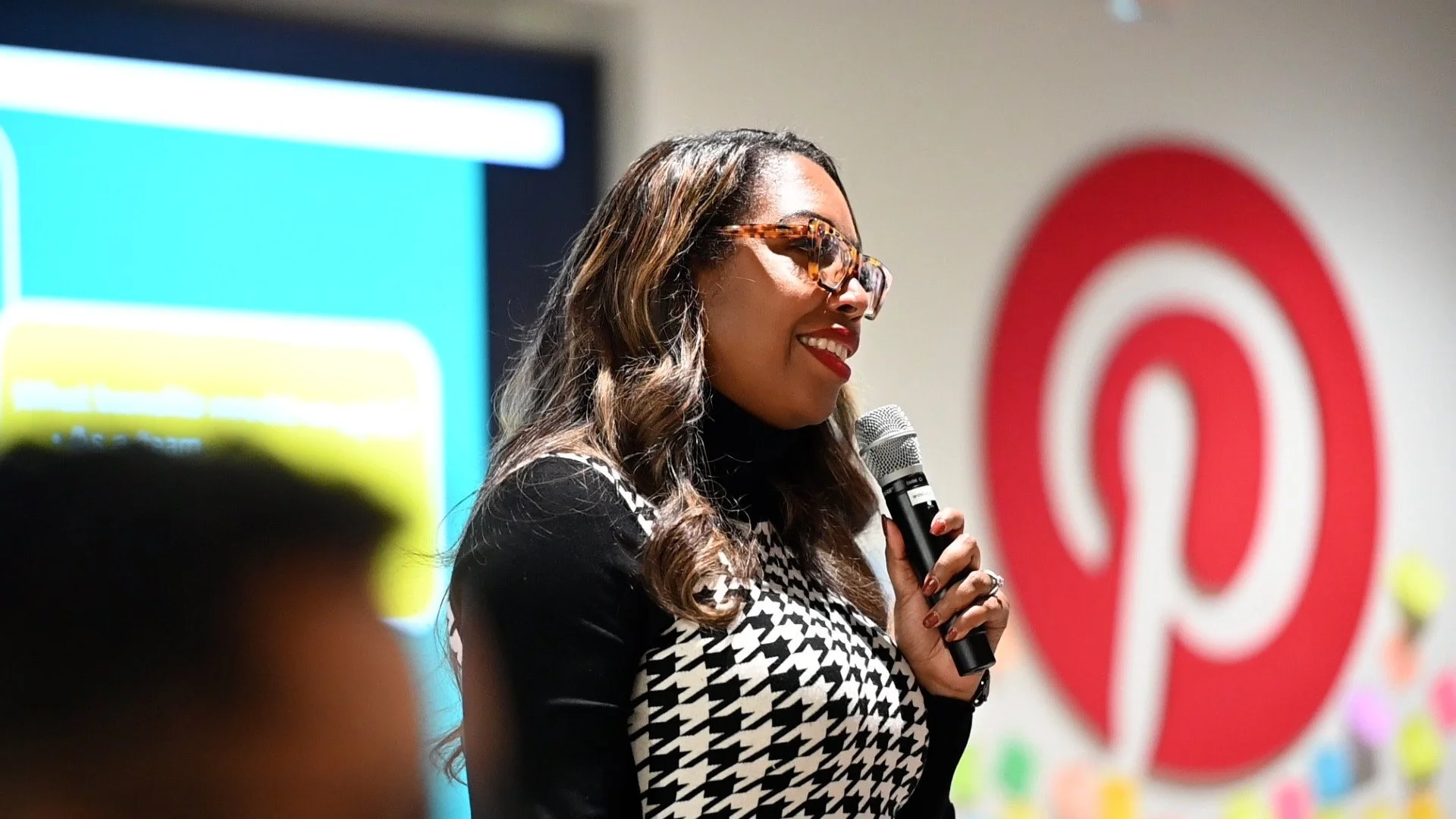 Woman in glasses speaking into a microphone at a presentation, with a large Pinterest logo on the wall behind her.