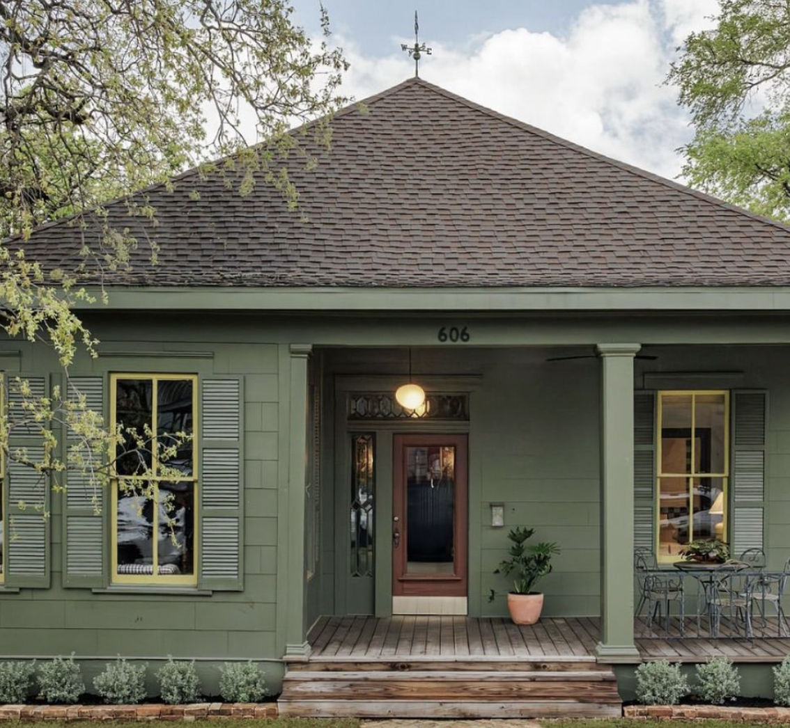 Front view of a green house with a brown shingled roof, yellow-trimmed windows, a wooden porch with potted plant, and a table with metal chairs.