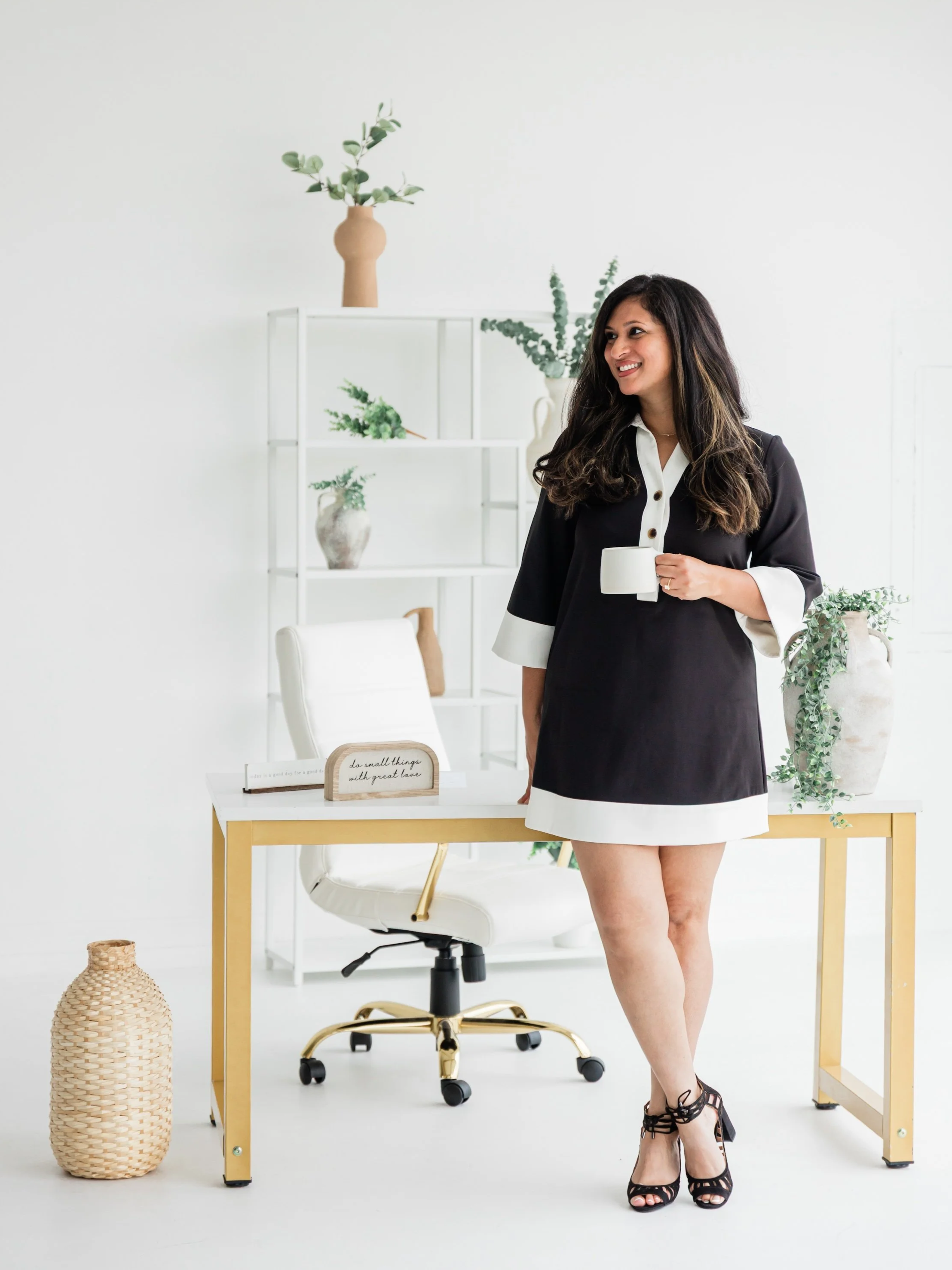 A woman in a black and white dress standing in a modern, minimalist office with white walls, holding a white mug, with a white desk, white chair, and decorative plants in pots.