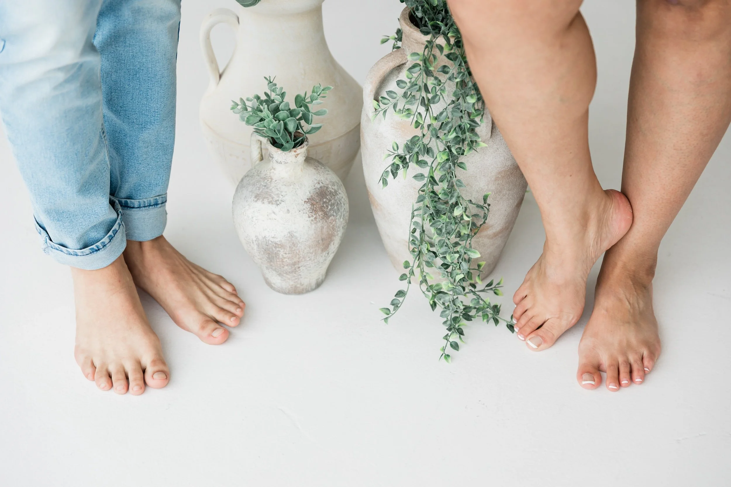 Two people standing barefoot on a white surface with three decorative vases and greenery in between.