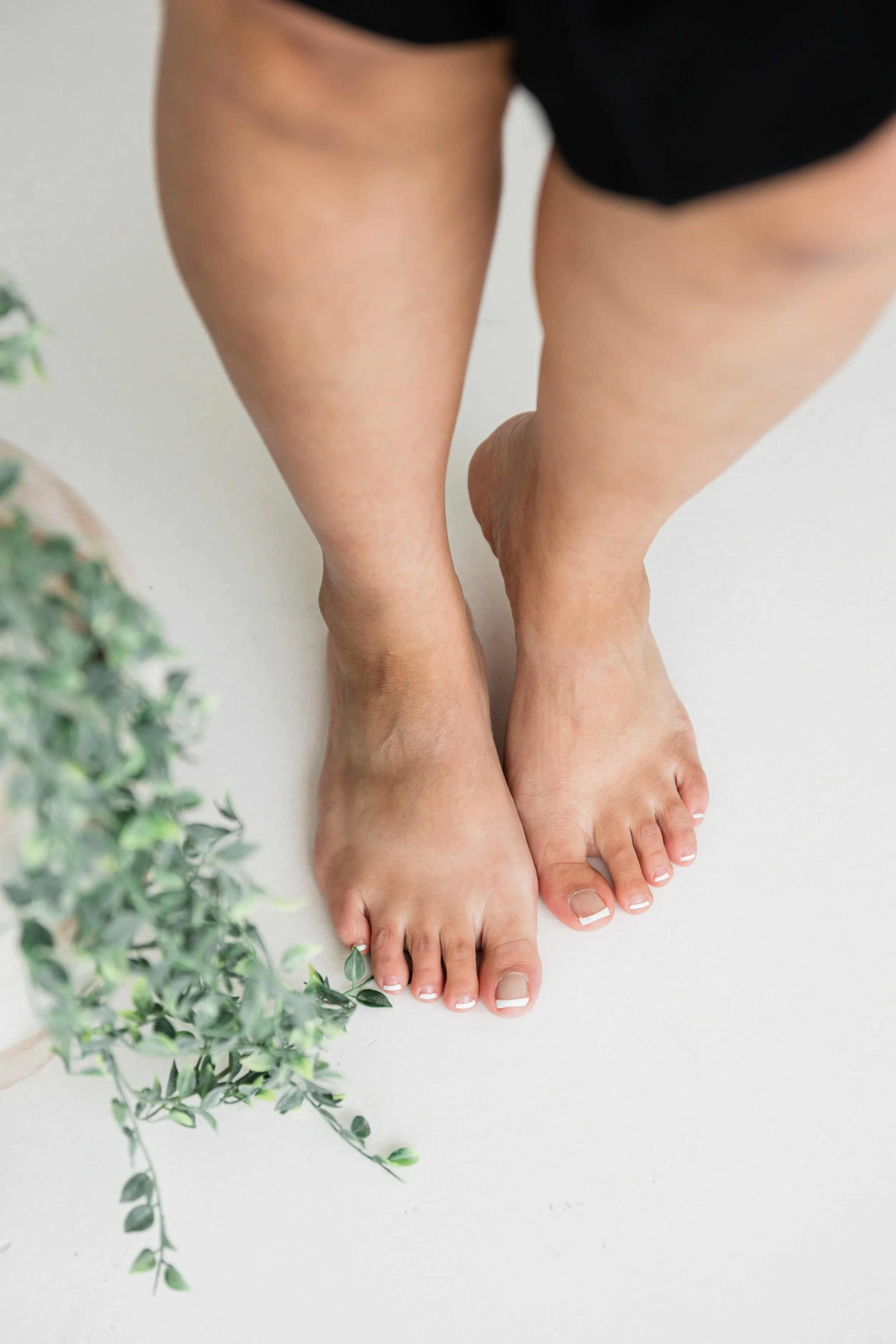 A person standing on a white floor, with part of their legs and feet visible, wearing black shorts. There are green plants in the foreground on the left side.