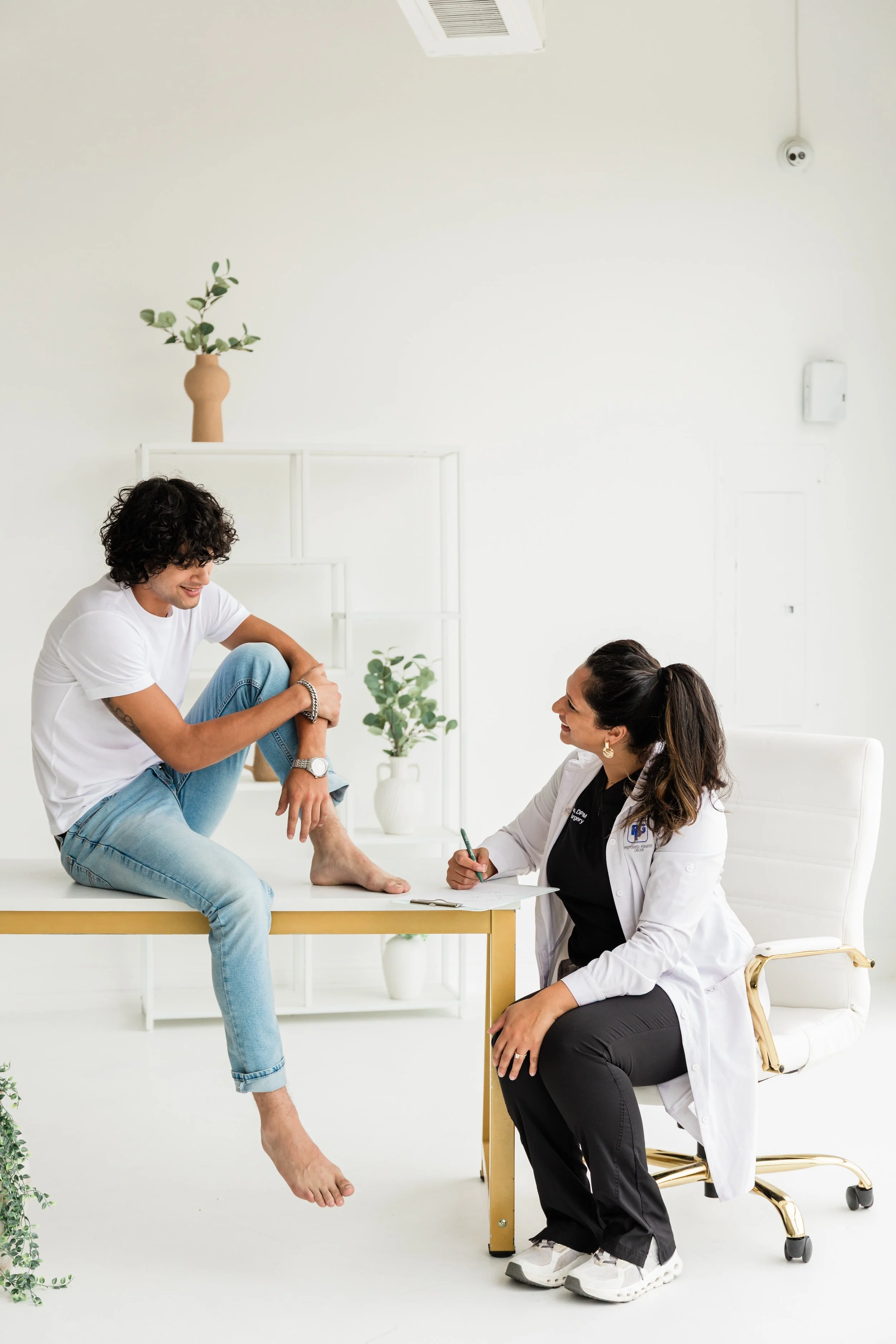 A doctor sitting at a desk taking notes while a patient sits on the desk, holding his ankle in a medical consultation room with white walls and a white chair.