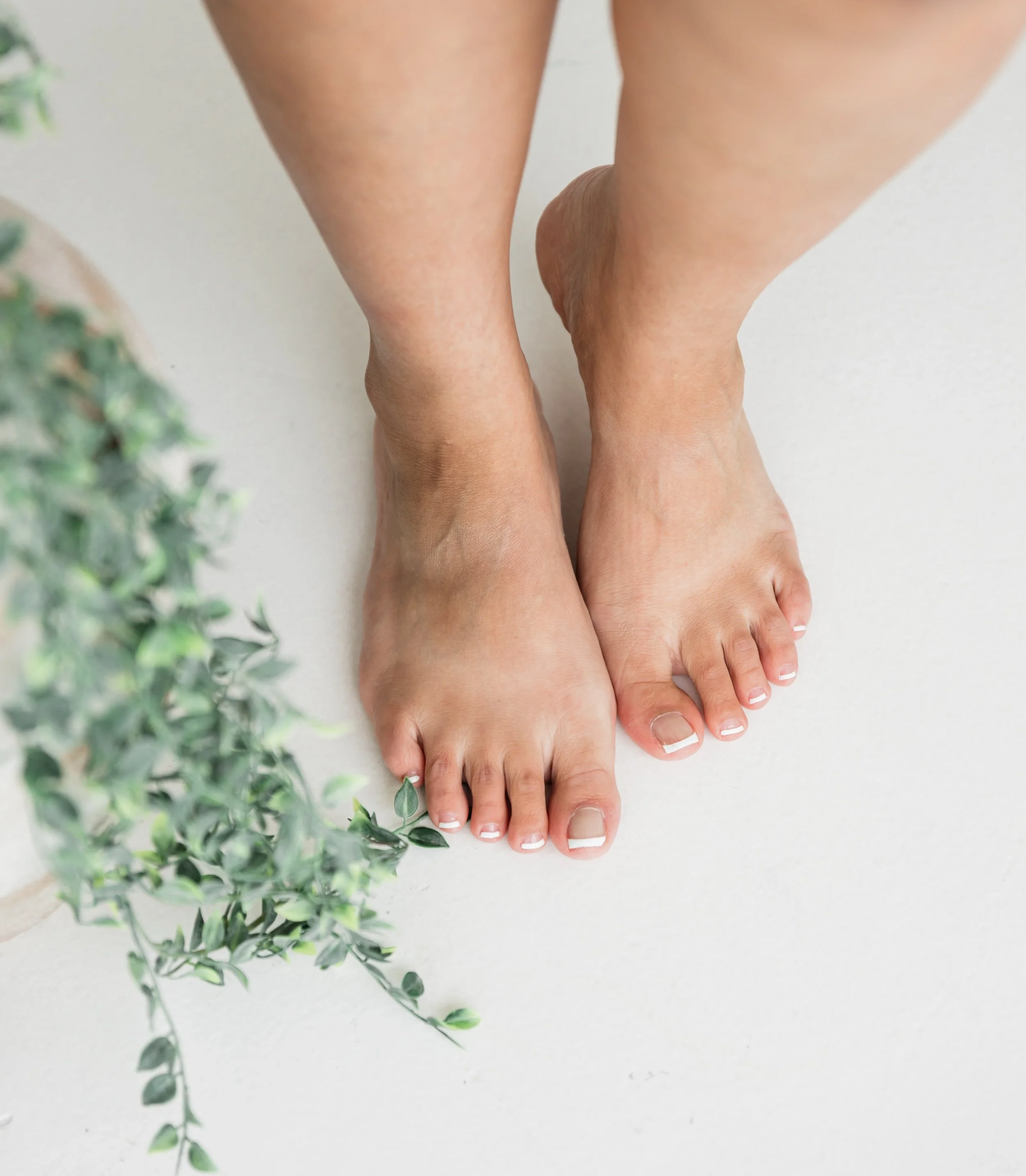 A person standing barefoot on a white floor with green leafy plant in the foreground.