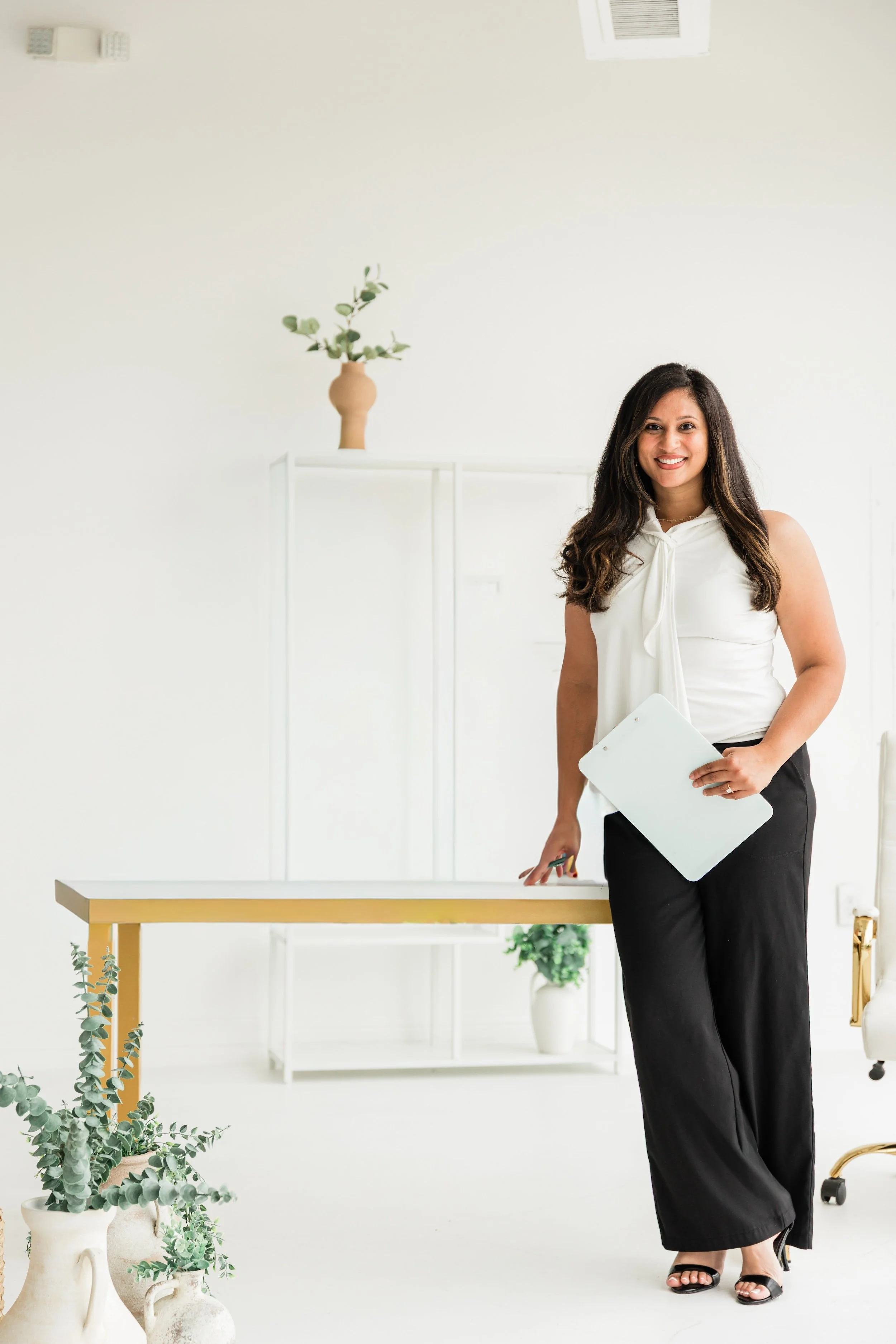 A woman standing in a bright, minimalist office holding a clipboard, smiling, with plants and white furniture in the background.