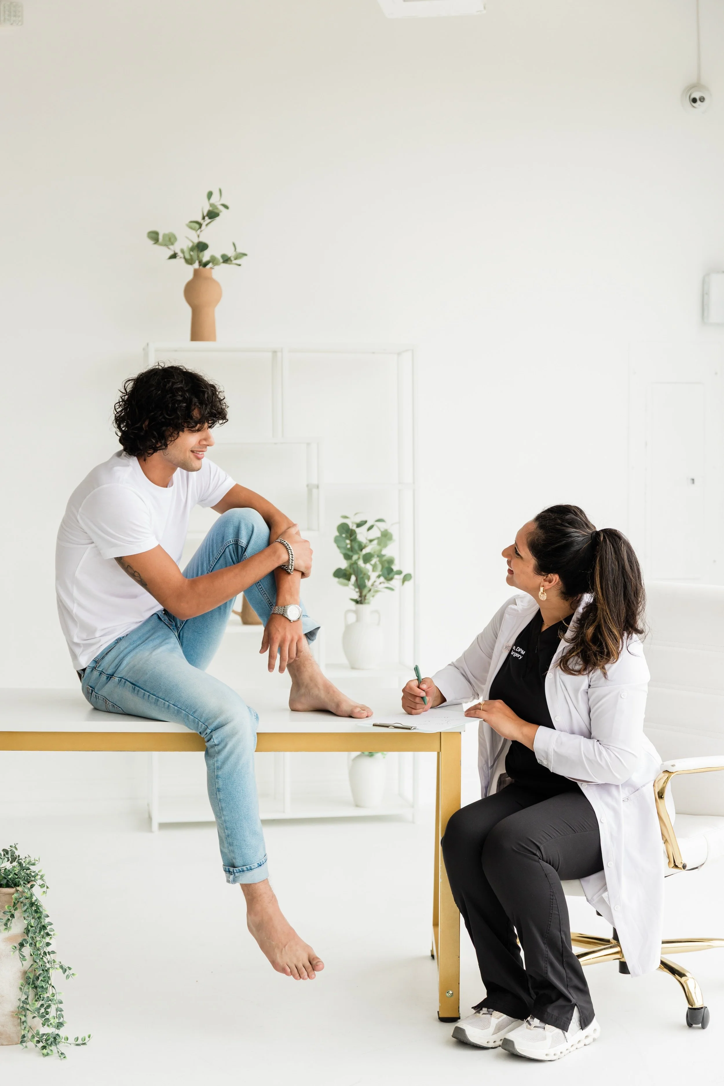 A young man sitting on a medical examination table with one leg raised and resting on the surface, talking to a female healthcare professional who is seated and taking notes.