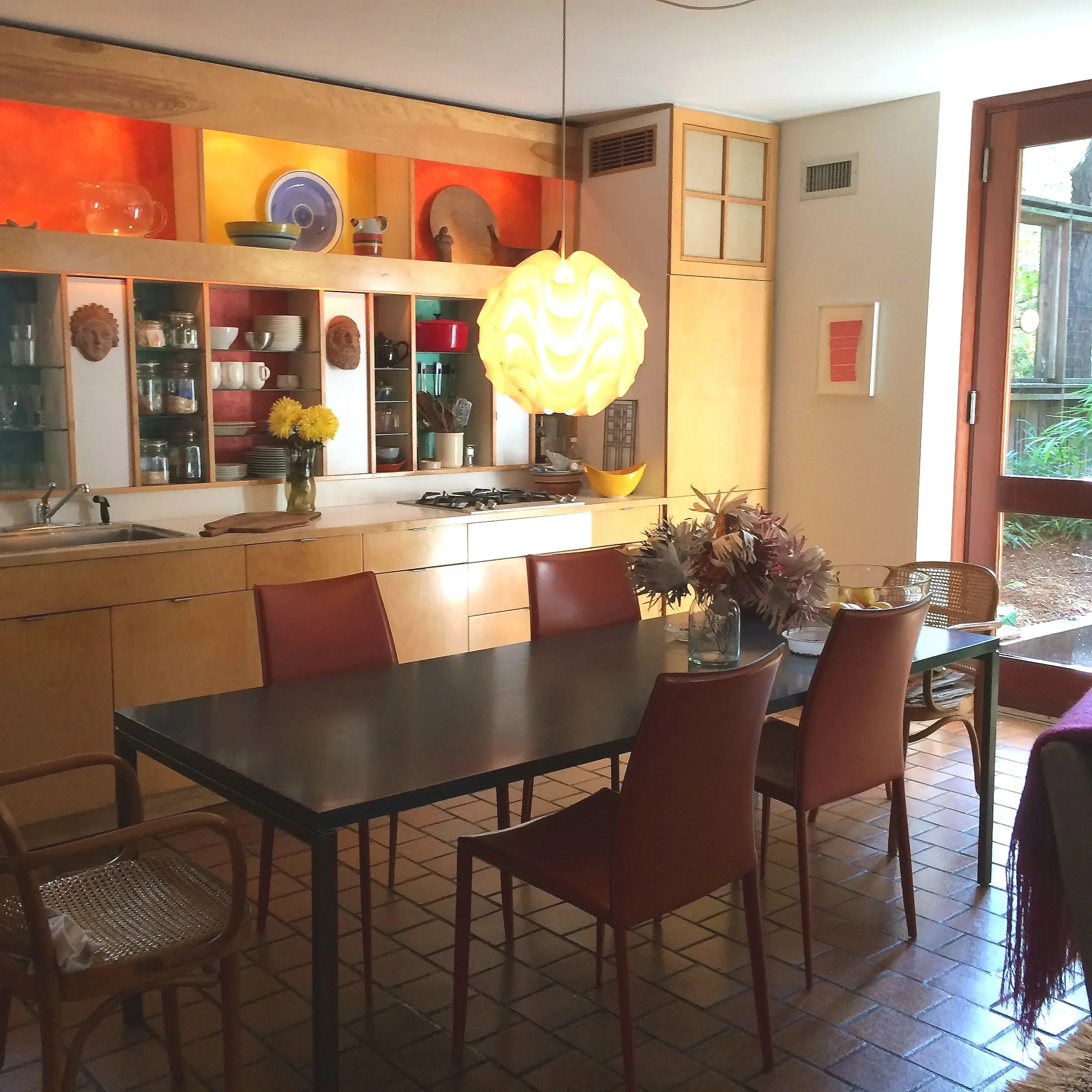 A cozy kitchen and dining area with a black table surrounded by a mix of chairs, a wooden cabinet with open shelves filled with dishes, colorful decorations, and a large window showing greenery outside. A distinctive yellow pendant lamp hangs above t